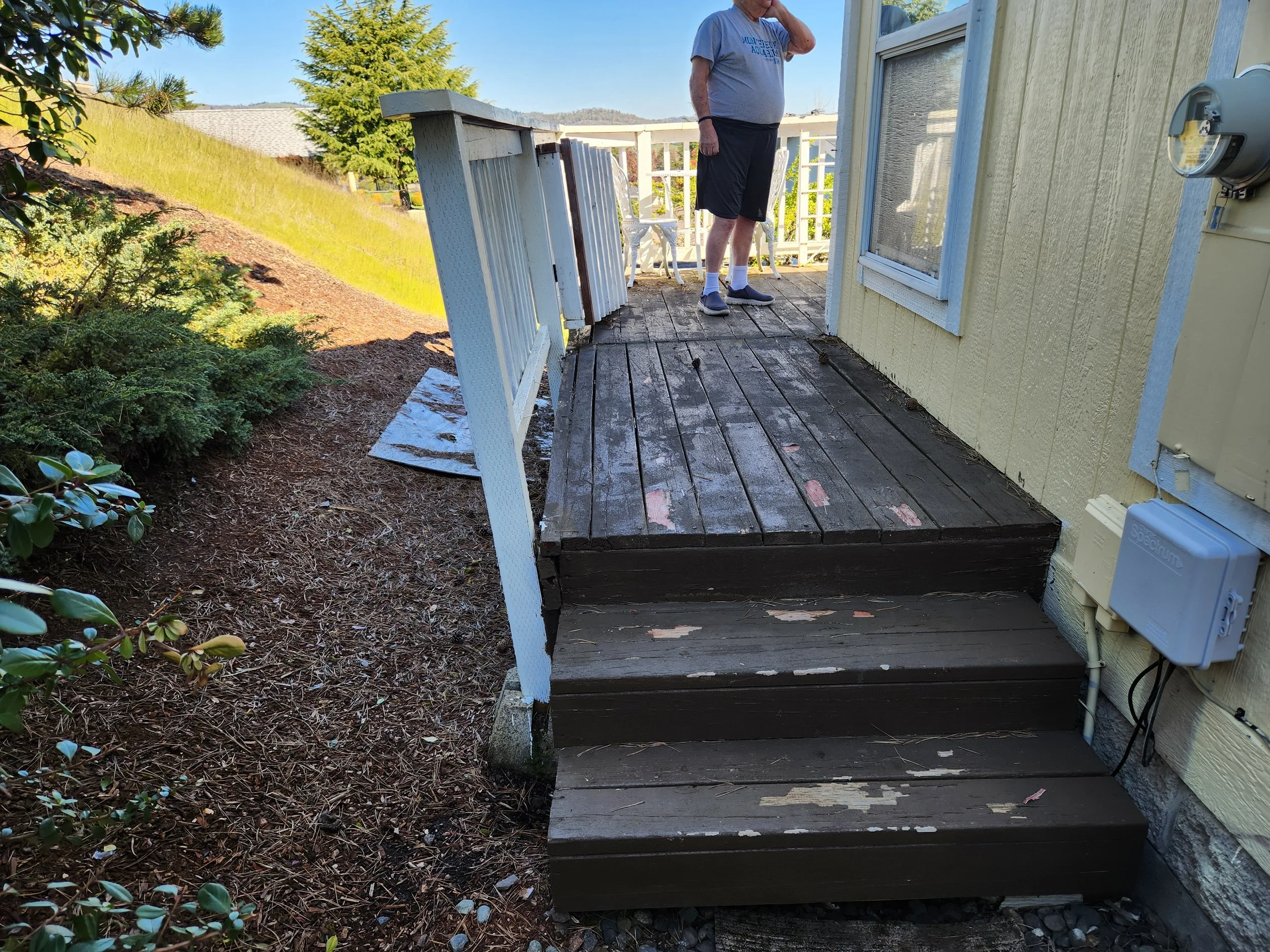 Weathered wooden porch with steps leading down, attached to a yellow house with a window. The porch is surrounded by plants and a garden, with a sloped grassy hill in the background under a clear blue sky.