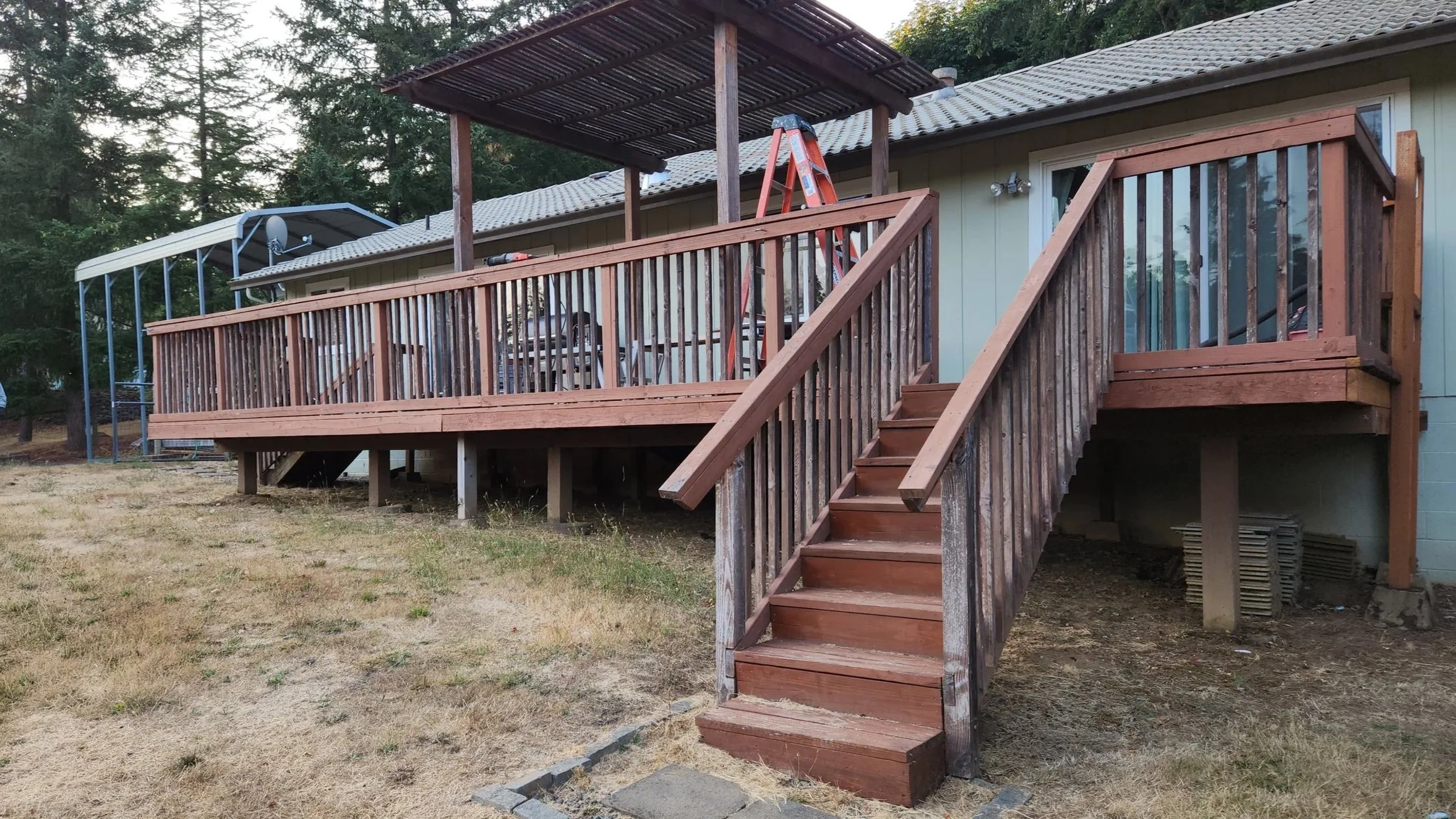 Before photo elevated wooden rotted  deck, stairs, and railing, with a grassy yard below and trees in the background.