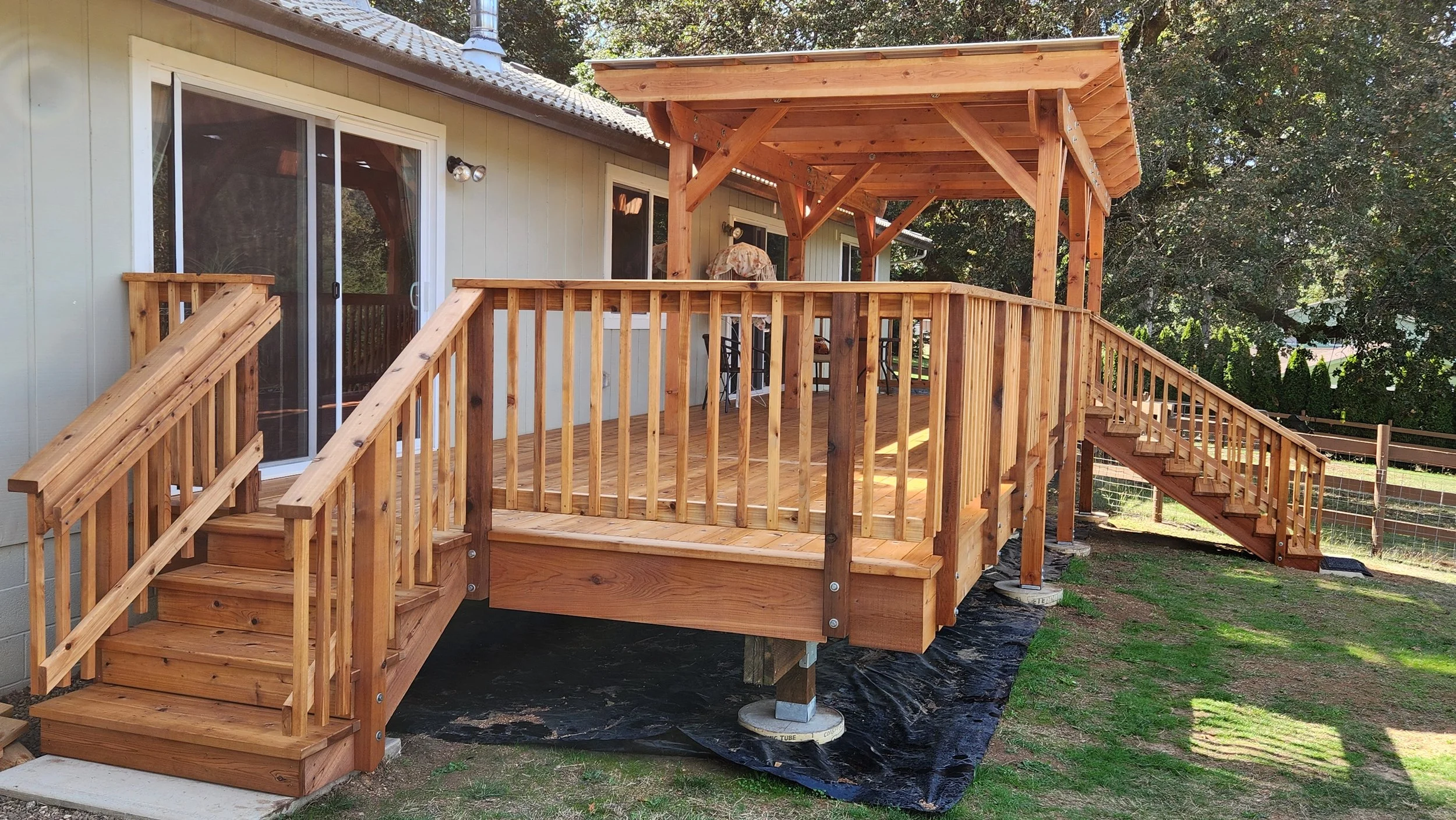Finished cedar deck with stairs leading to a house, supported by concrete and pressure treated posts, with a privacy fence on the right, and a covered section with a gazebo/pergola on top.
