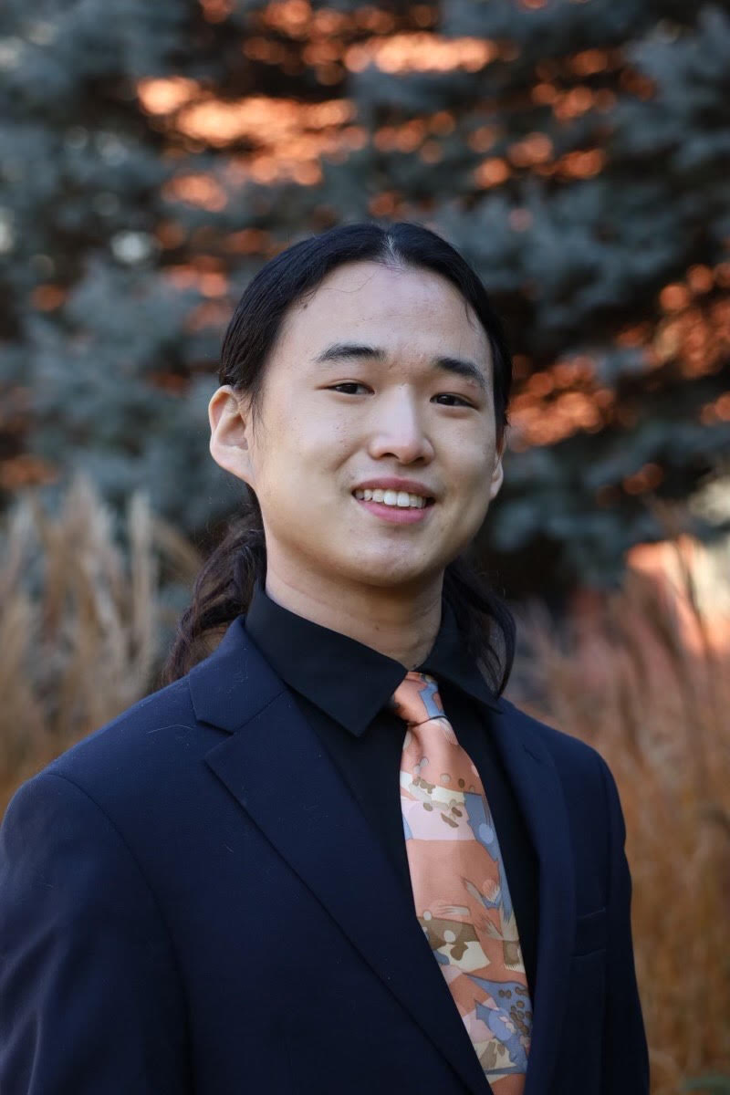 A young man with long black hair, wearing a dark suit and a colorful patterned tie, standing outdoors with a blurred background of trees and foliage during sunset.