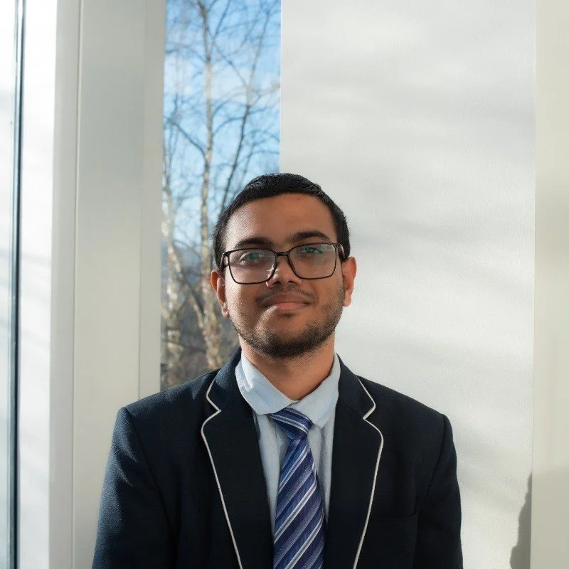 A young man with glasses, dressed in a dark blazer with piping, white shirt, and striped tie, standing indoors near a window with a view of leafless trees outside.