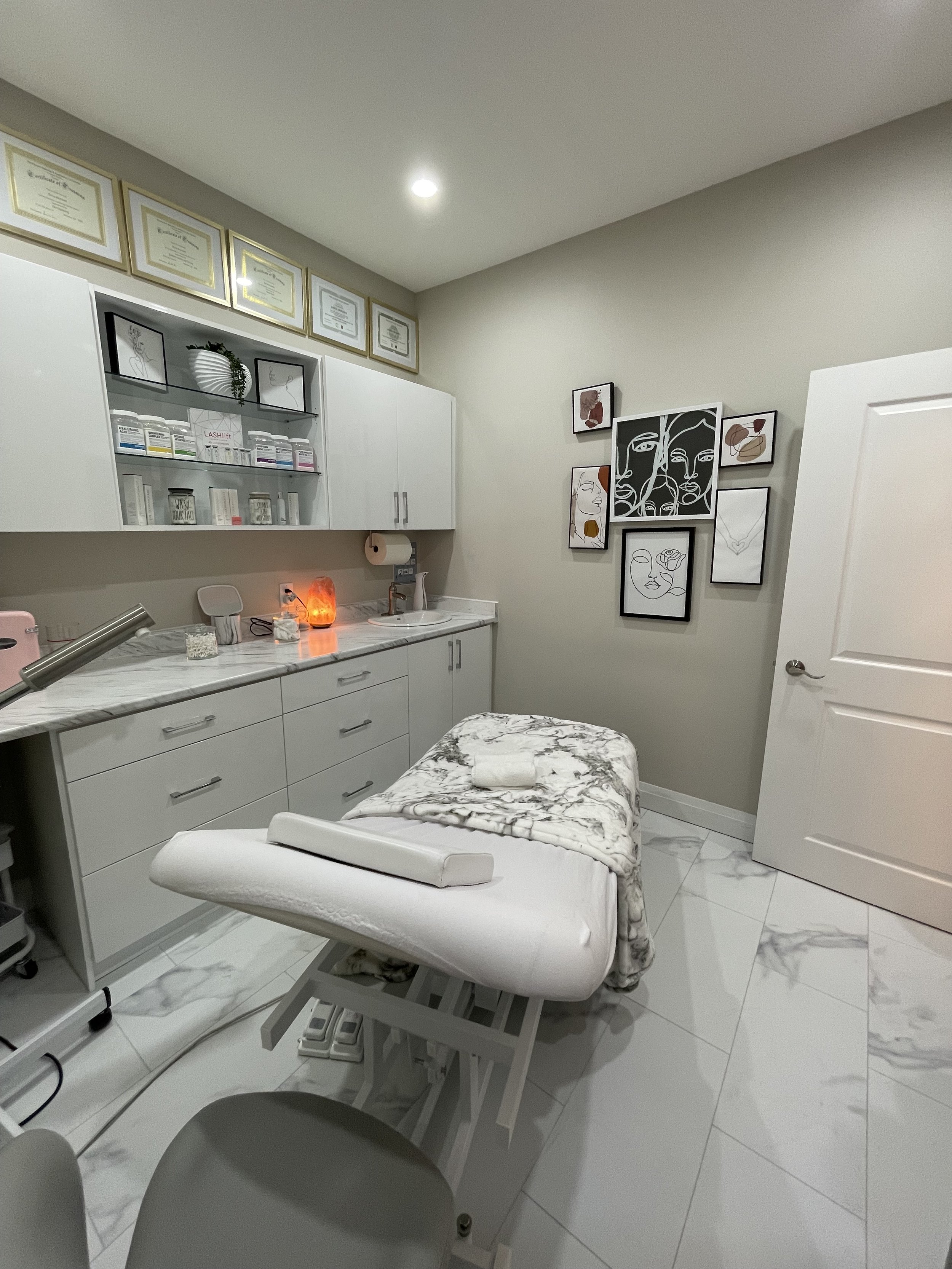 A medical or treatment room with a massage or examination table, white cabinets, framed certificates, wall art, and a salt lamp.
