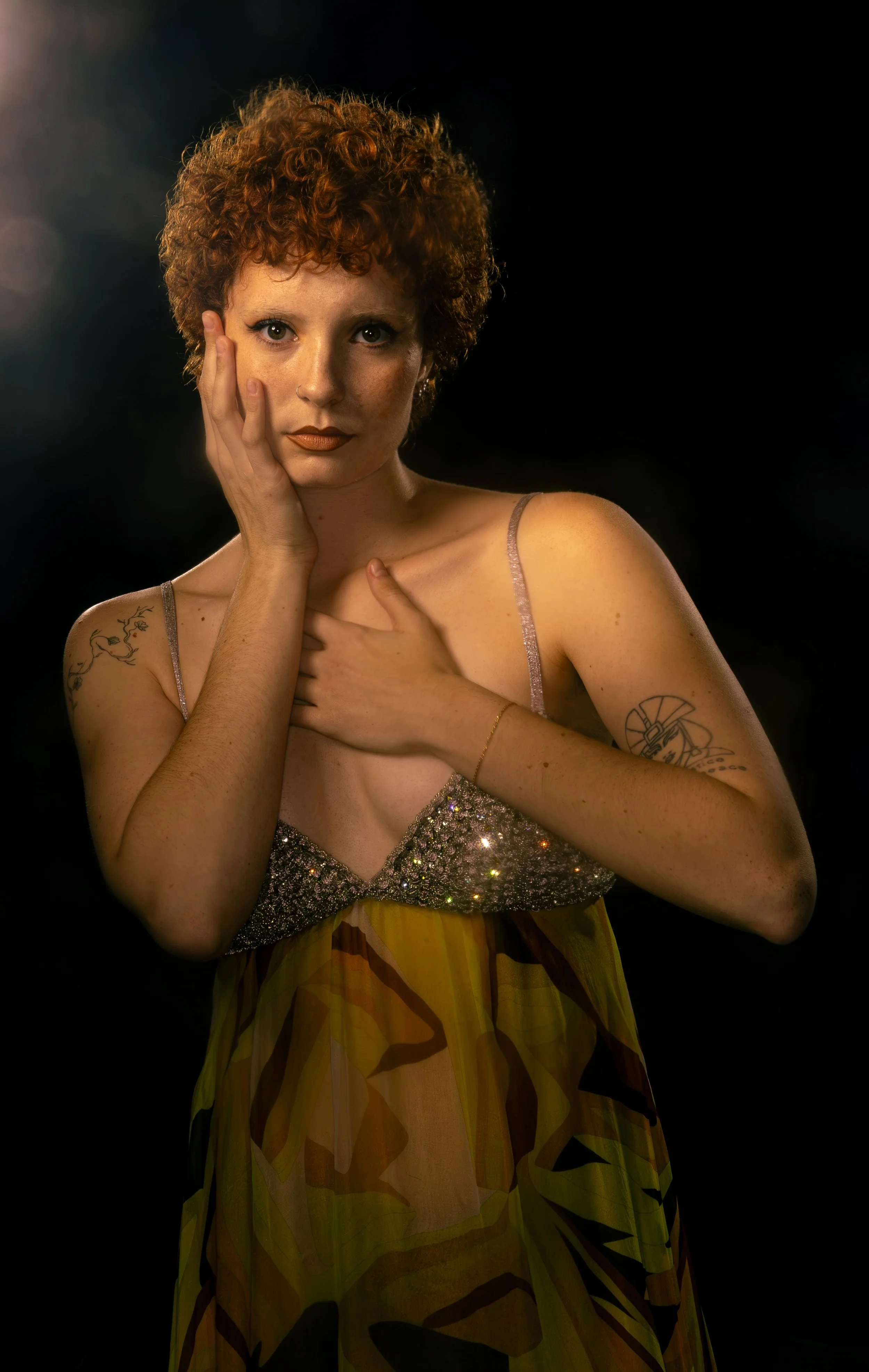 Portrait of a woman with short curly red hair posing against a black background, wearing a spaghetti strap top with glittery details and a colorful, abstract patterned skirt, with tattoos on both arms, touching her face and chest.