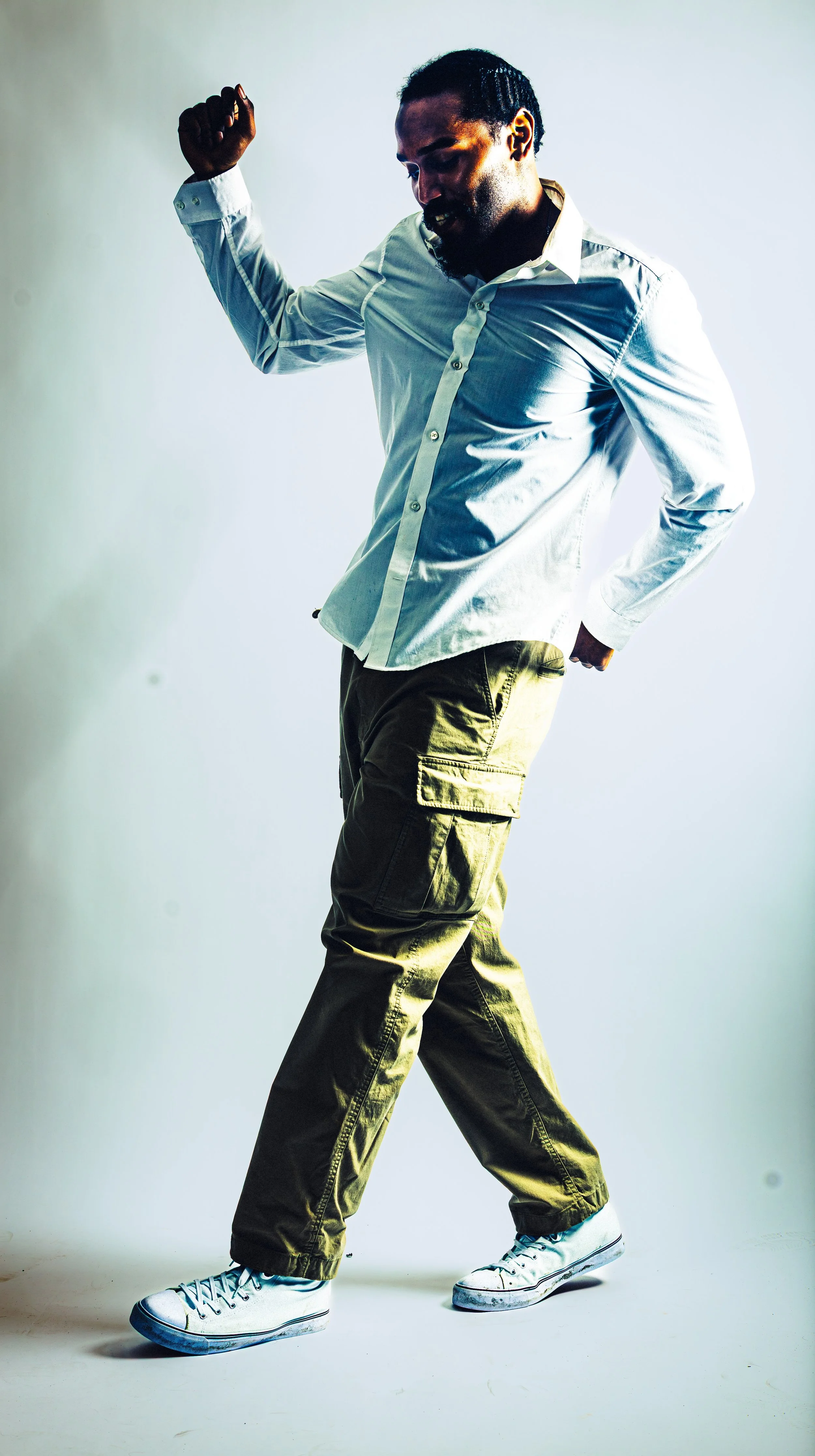A man dancing, wearing a white shirt, cargo pants, and sneakers, with one arm raised and looking down, in a studio setting.