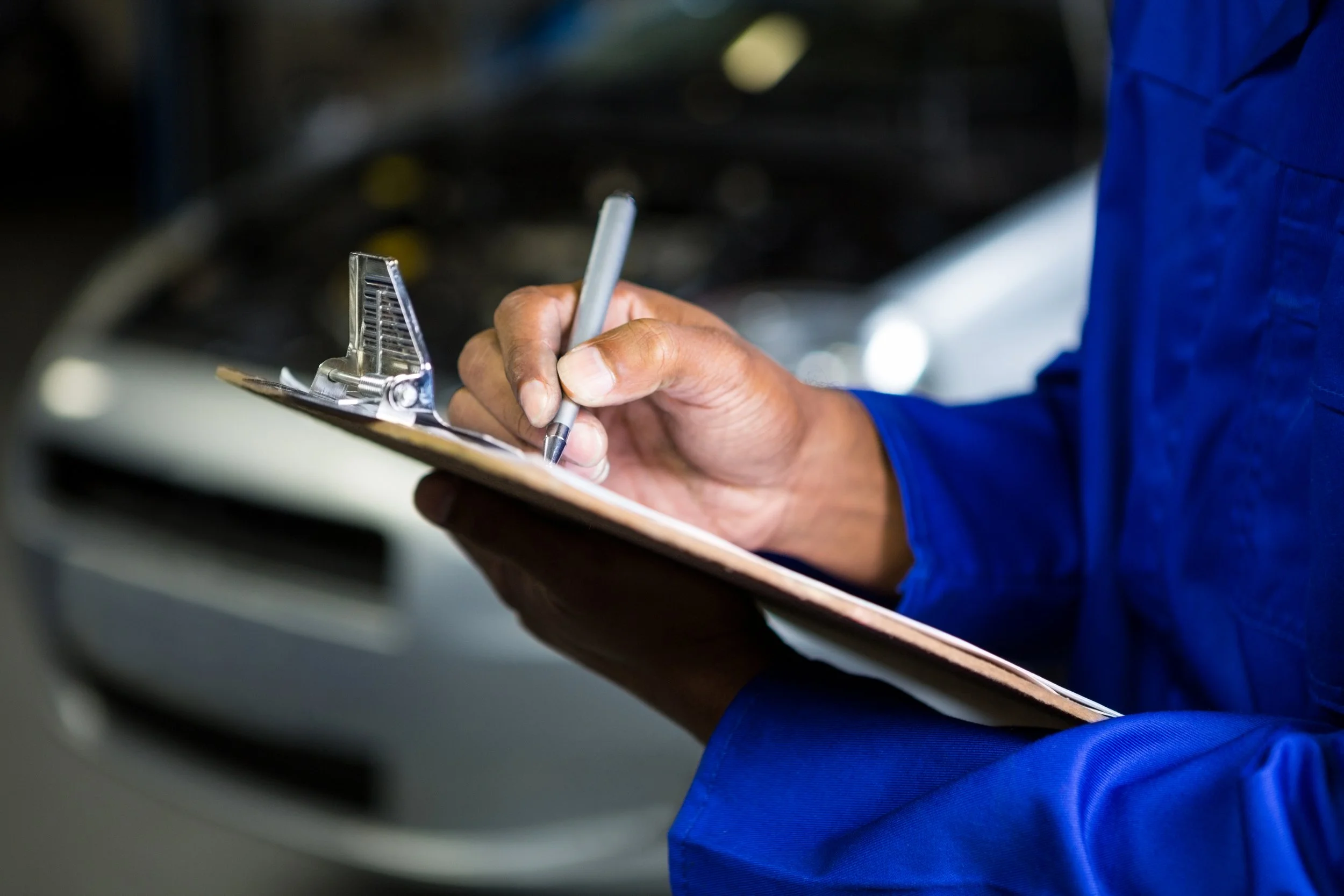 A person in blue overalls writes on a form with a pen, with a car door in the background.