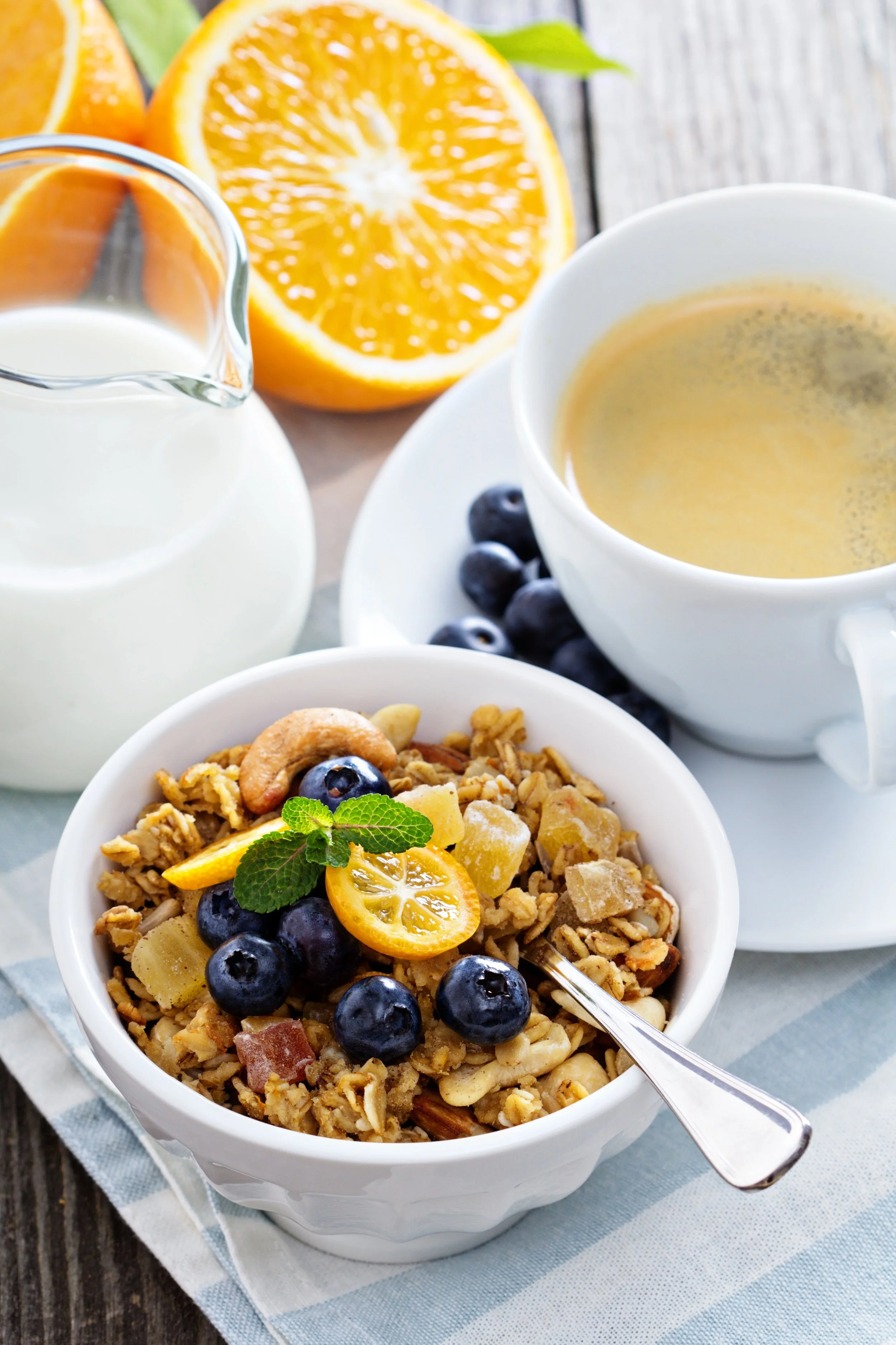 Frokost med granola med blåbær og sitrusskiver, en kopp kaffe, et glass melk og en halveret appelsin på en treplate.