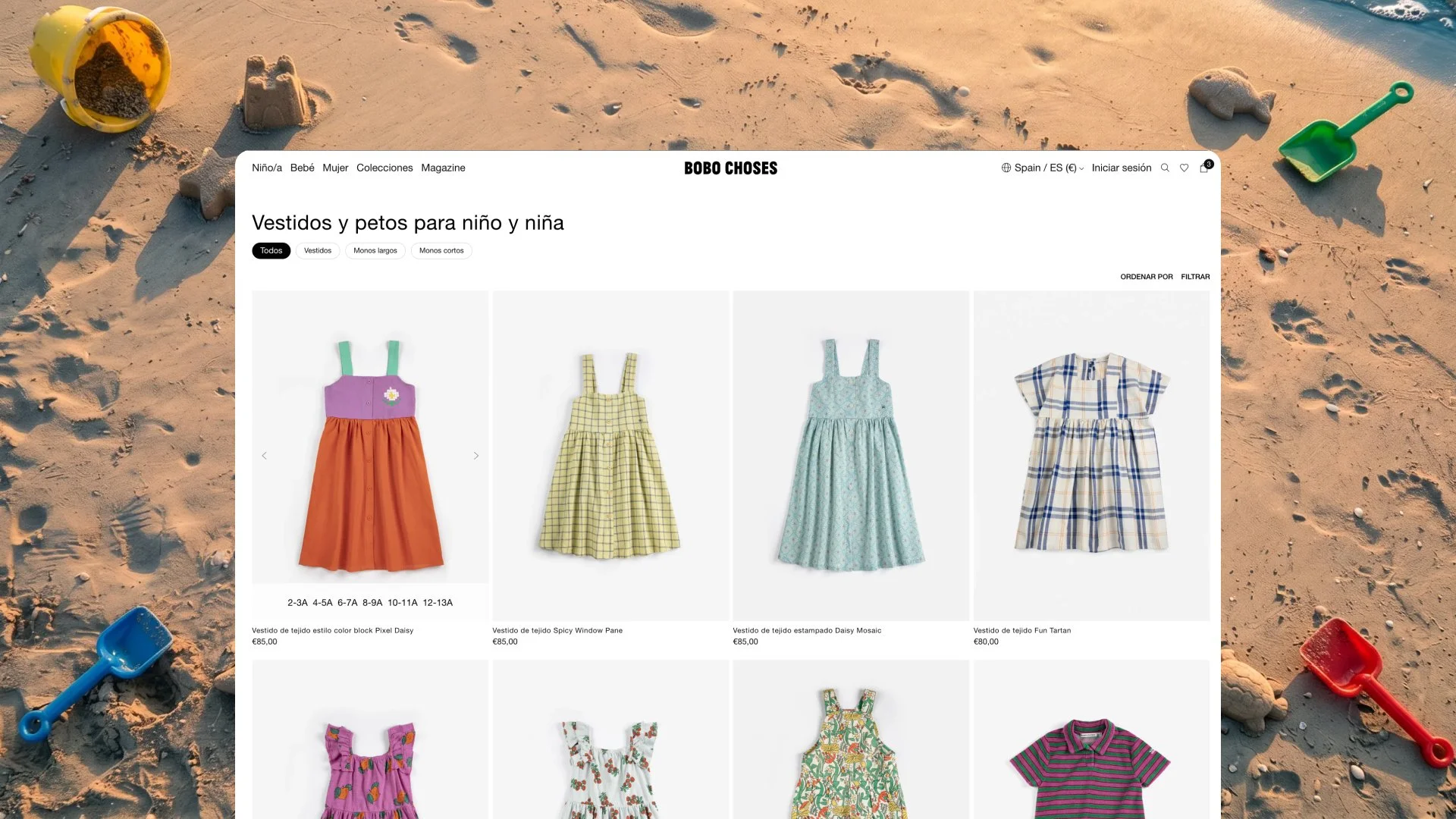 Página de tienda online Bobo Choses con catálogo de vestidos y petos infantiles, mostrada sobre fondo de arena de playa con palas y moldes de juguete.