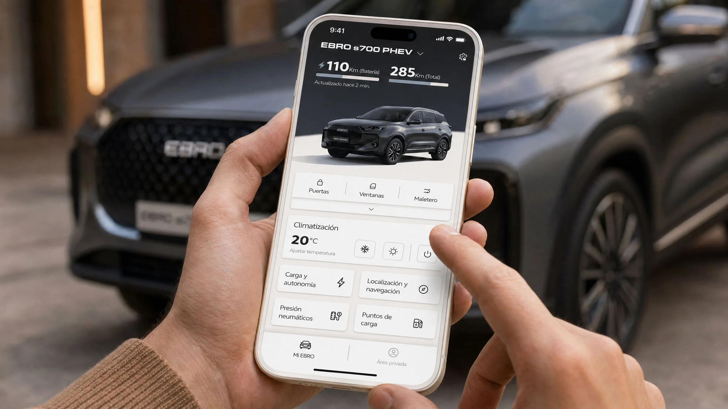 Persona sostiene un smartphone con aplicación de control del vehículo eléctrico Ebro s700 PHEV, mostrando autonomía, climatización y funciones remotas, con el coche al fondo.