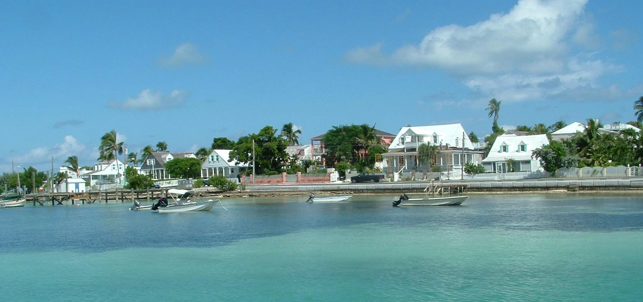 Turtle Bay Eleuthera Bahamas homes and building lots for sale. View of houses along a waterfront with boats docked at a pier under a blue sky with some clouds.