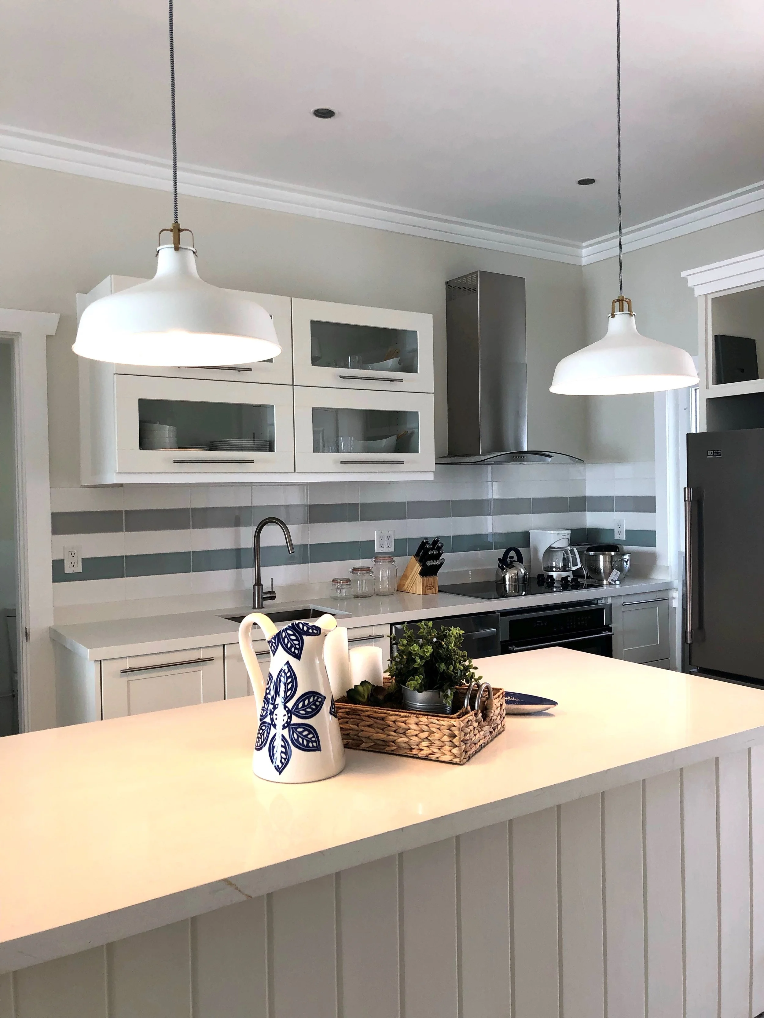 Turtle Bay Eleuthera Bahamas homes andModern kitchen with white cabinets, grey backsplash, stainless steel appliances, and two hanging pendant lights over a white island. On the island, there is a decorative pitcher, a potted plant, and a woven tray.