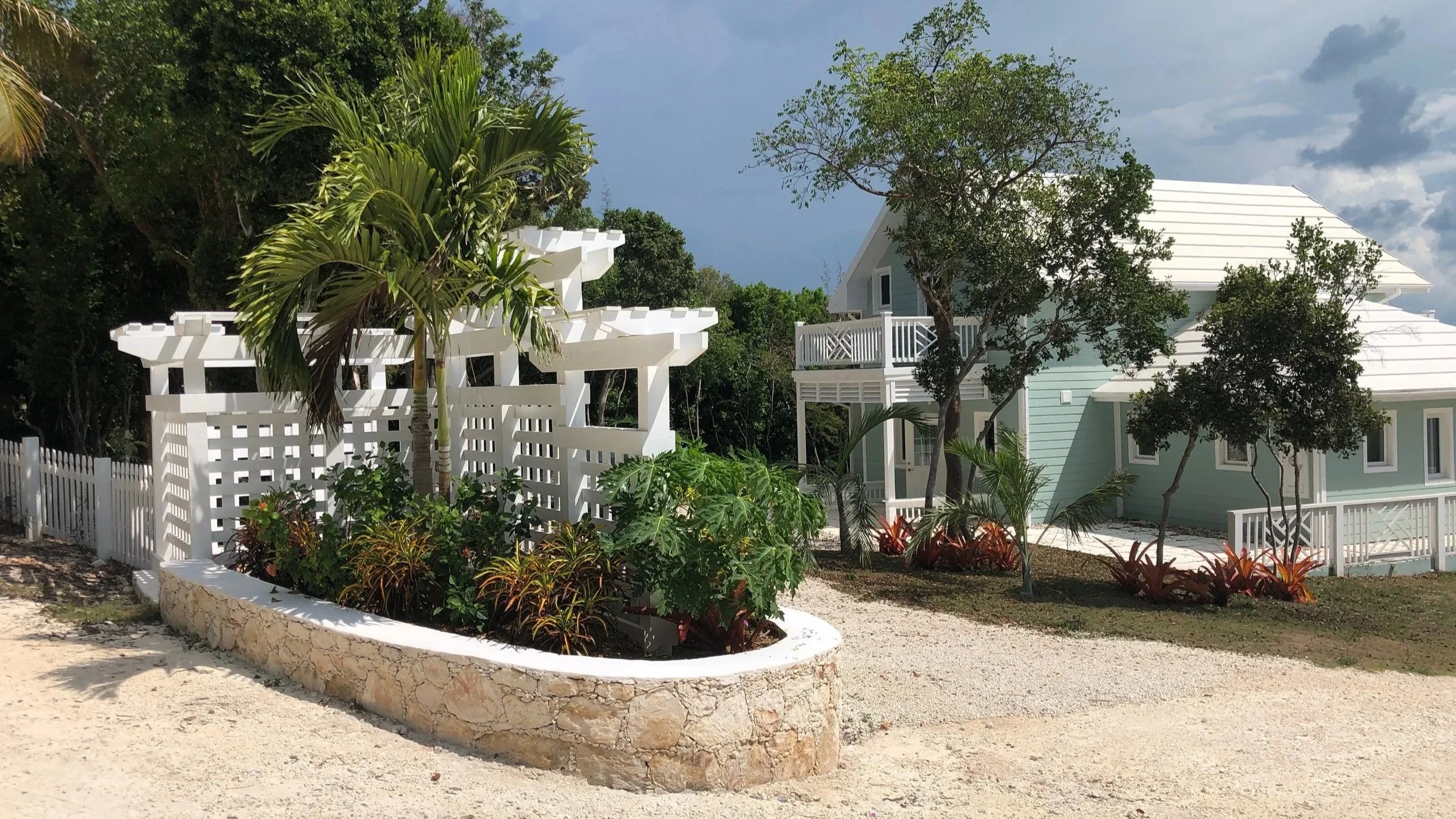 Turtle Bay Eleuthera Bahamas homes and building lots for sale. A light blue house with white trim, surrounded by trees and plants, sunny sky with some clouds, gravel driveway, and a white fence.