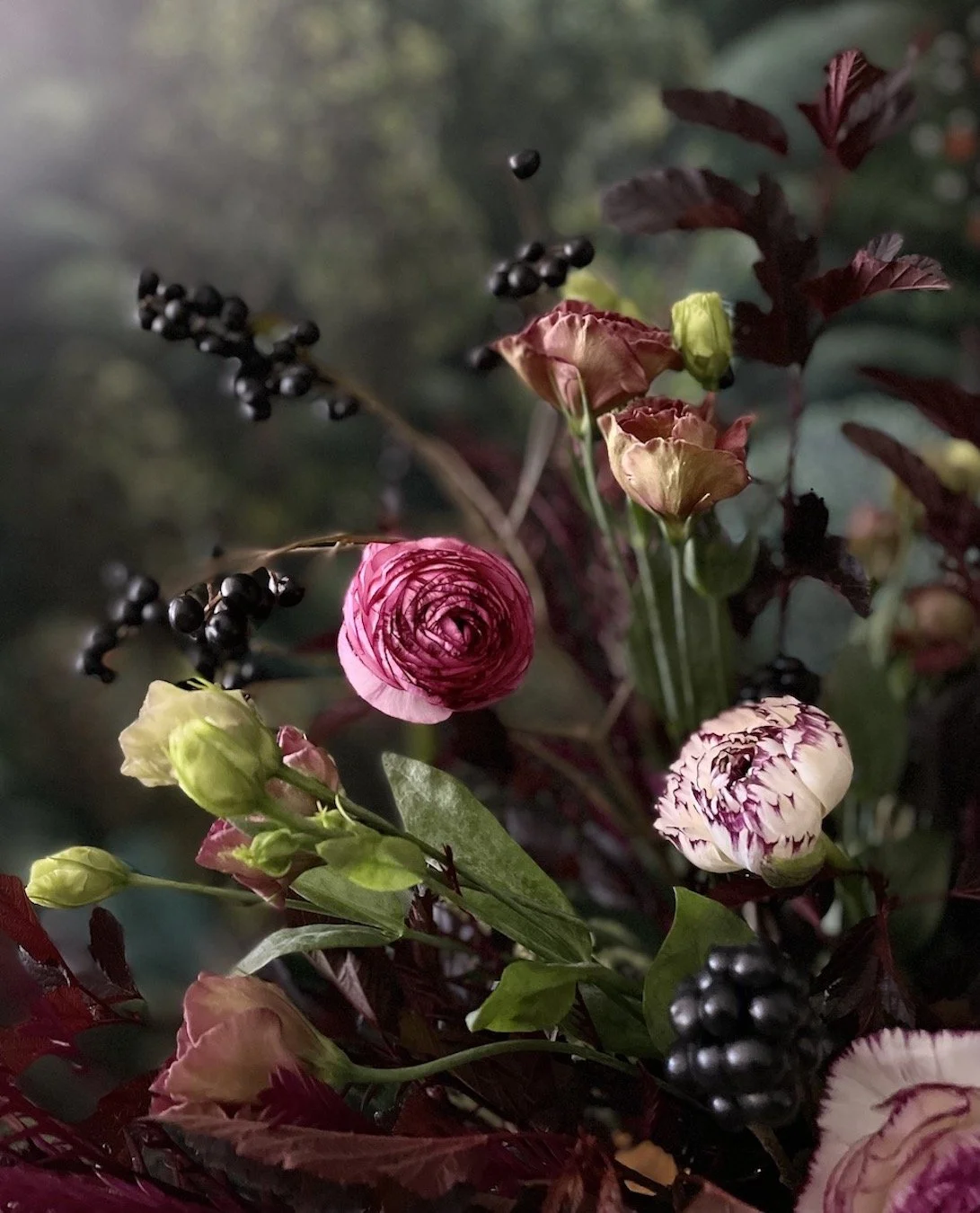 Arrangement of pink and white ranunculus flowers with green buds, dark purple berries, and leafy foliage.