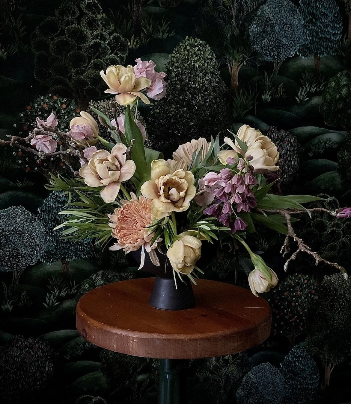 Flower arrangement with roses, tulips, and other flowers in a black vase on a wooden table, with a dark leafy background.