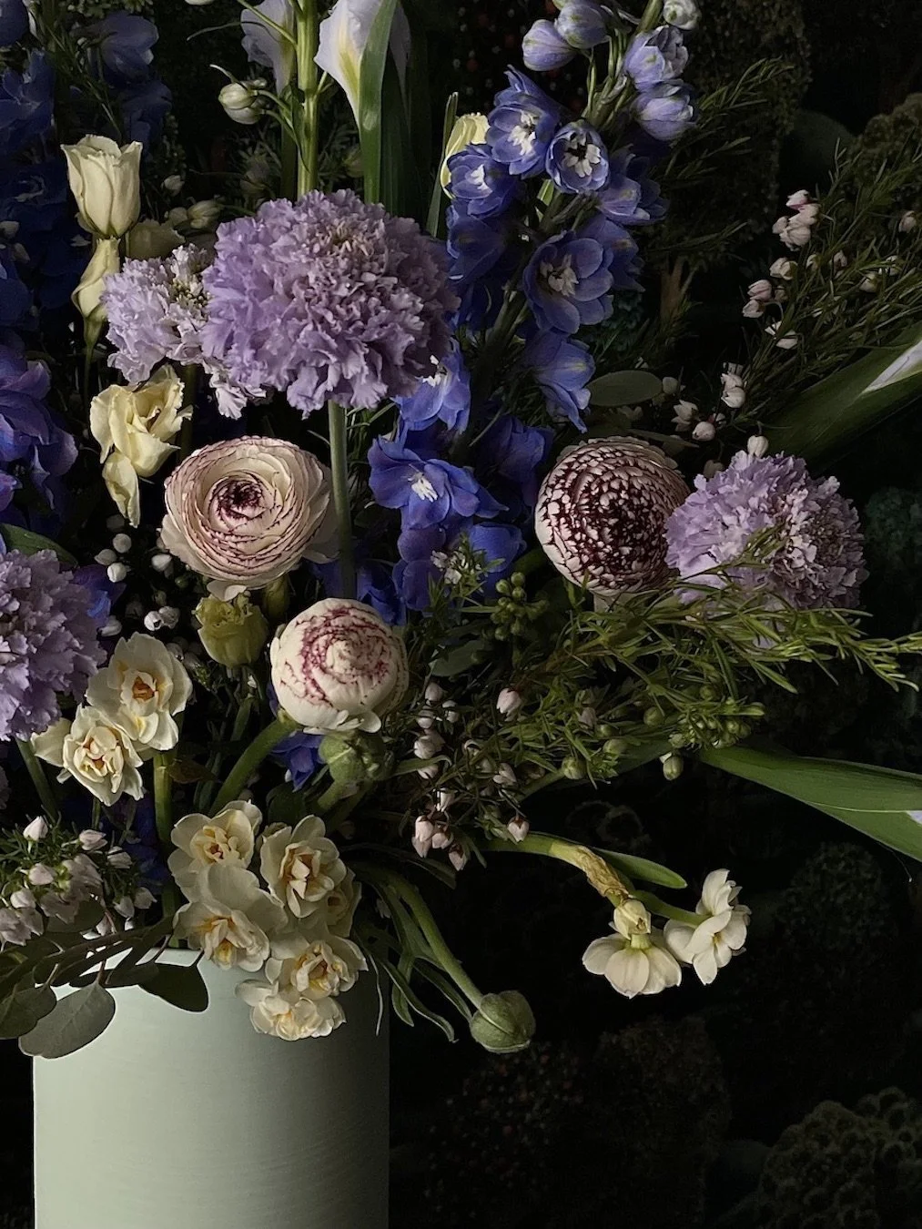 Close-up of a mixed floral bouquet in a white vase, featuring purple, white, and blue flowers with green foliage.