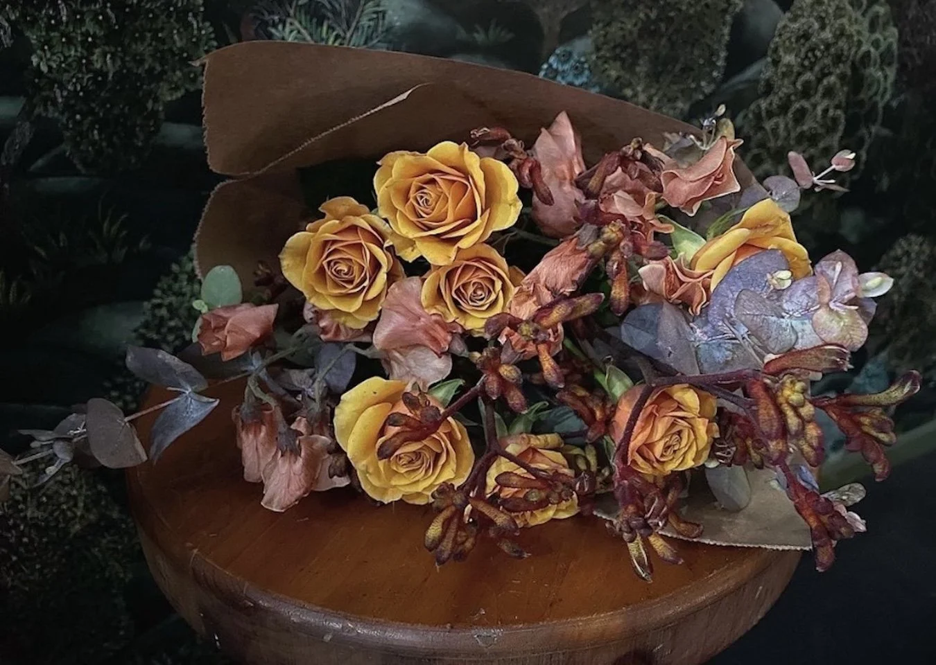 A bouquet of yellow roses, dried pink flowers, and purple leaves on a round wooden surface.