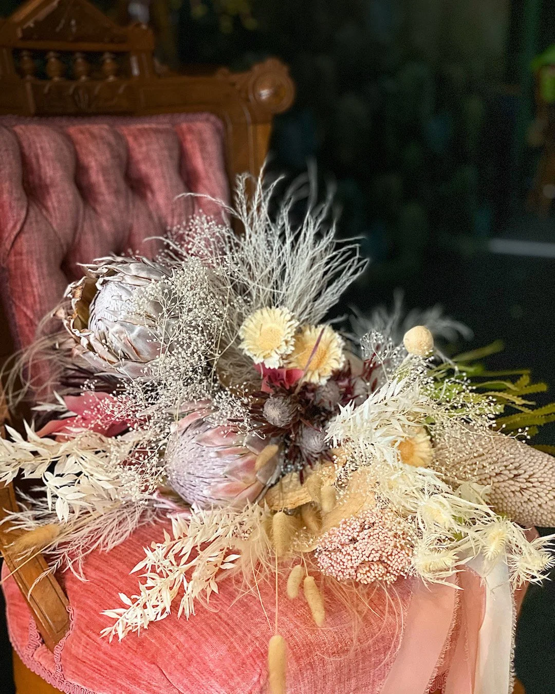 A bouquet of dried flowers and grasses on a pink fabric surface, with a carved wooden chair in the background.
