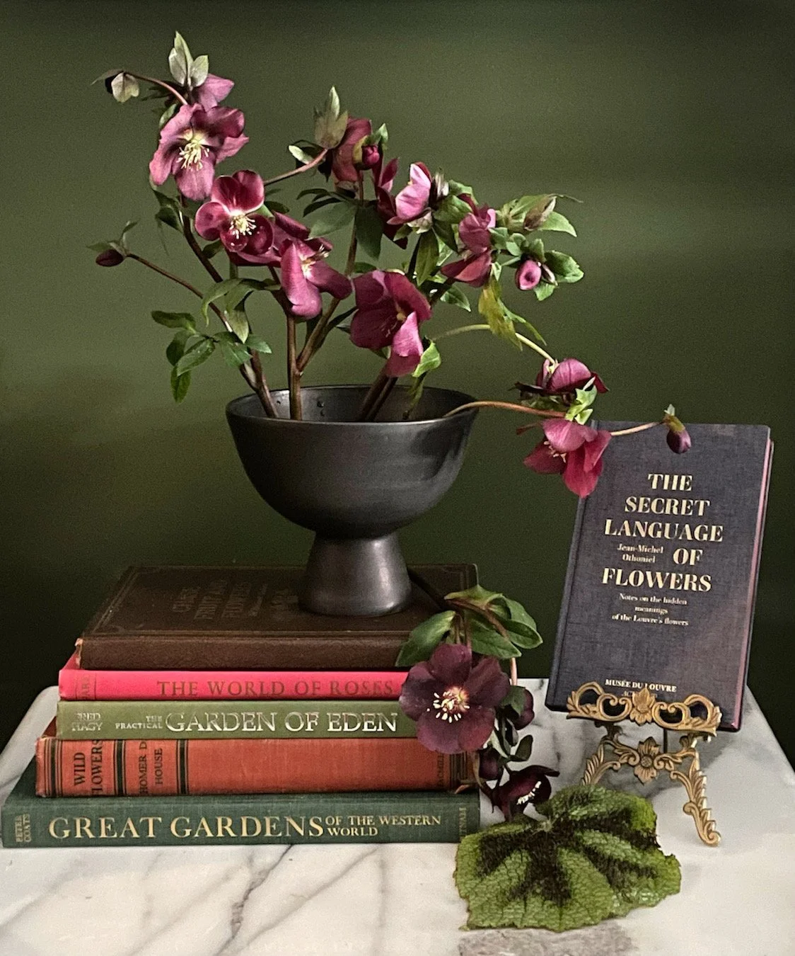 A black bowl with pink and purple flowers and green leaves, placed on top of three stacked gardening books, with a metal stand holding a book titled "The Secret Language of Flowers" on a marble surface against a green background.