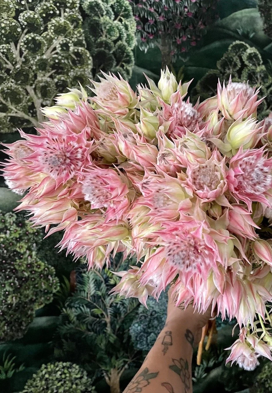 A person holding a large bouquet of pink and cream flowers with spiky petals, possibly artificial, against a background of various green plants and floral arrangements.