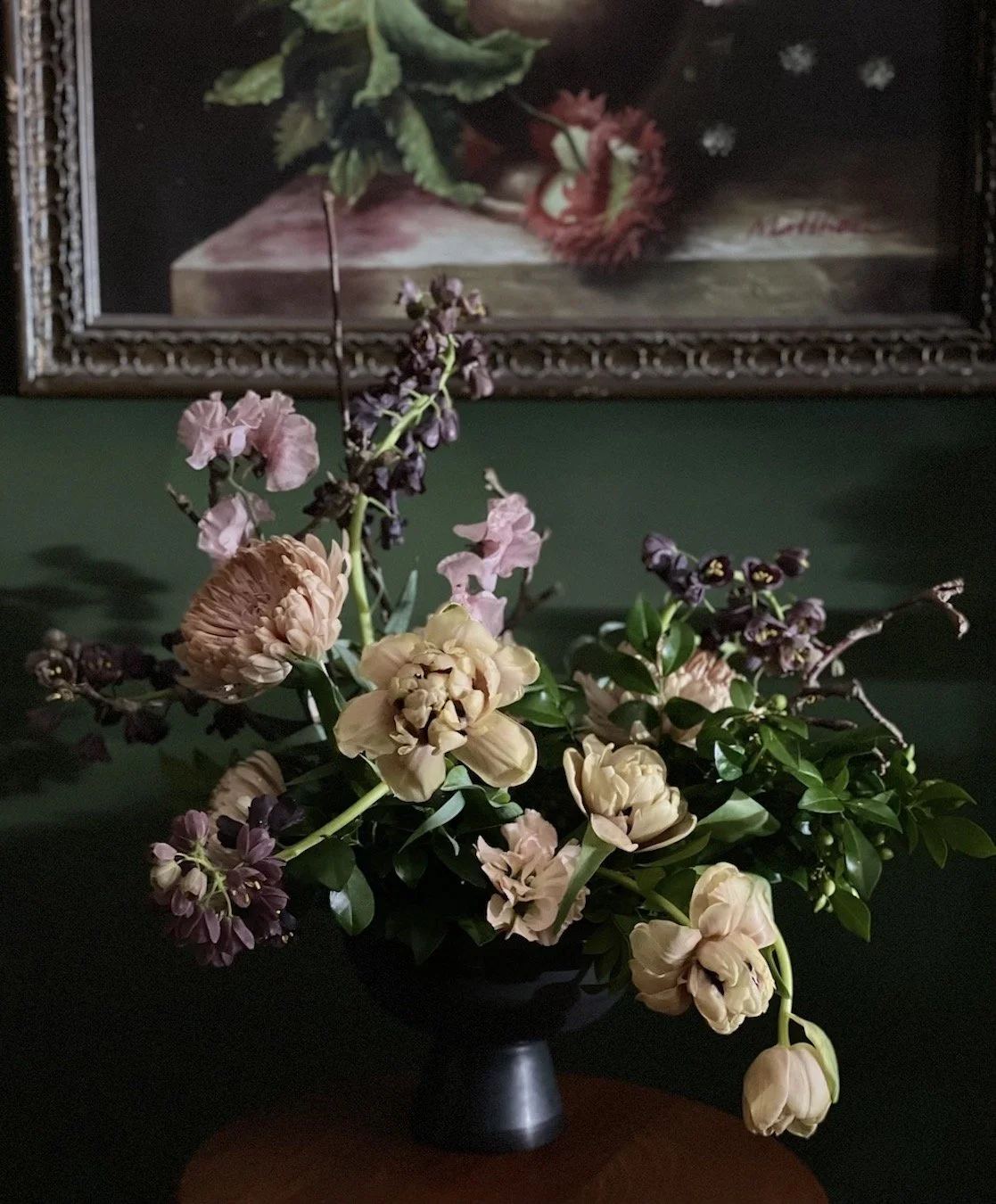 A floral arrangement with cream, purple, and pink flowers and green leaves in a black vase, set on a wooden table against a dark green wall with a framed still-life painting in the background.