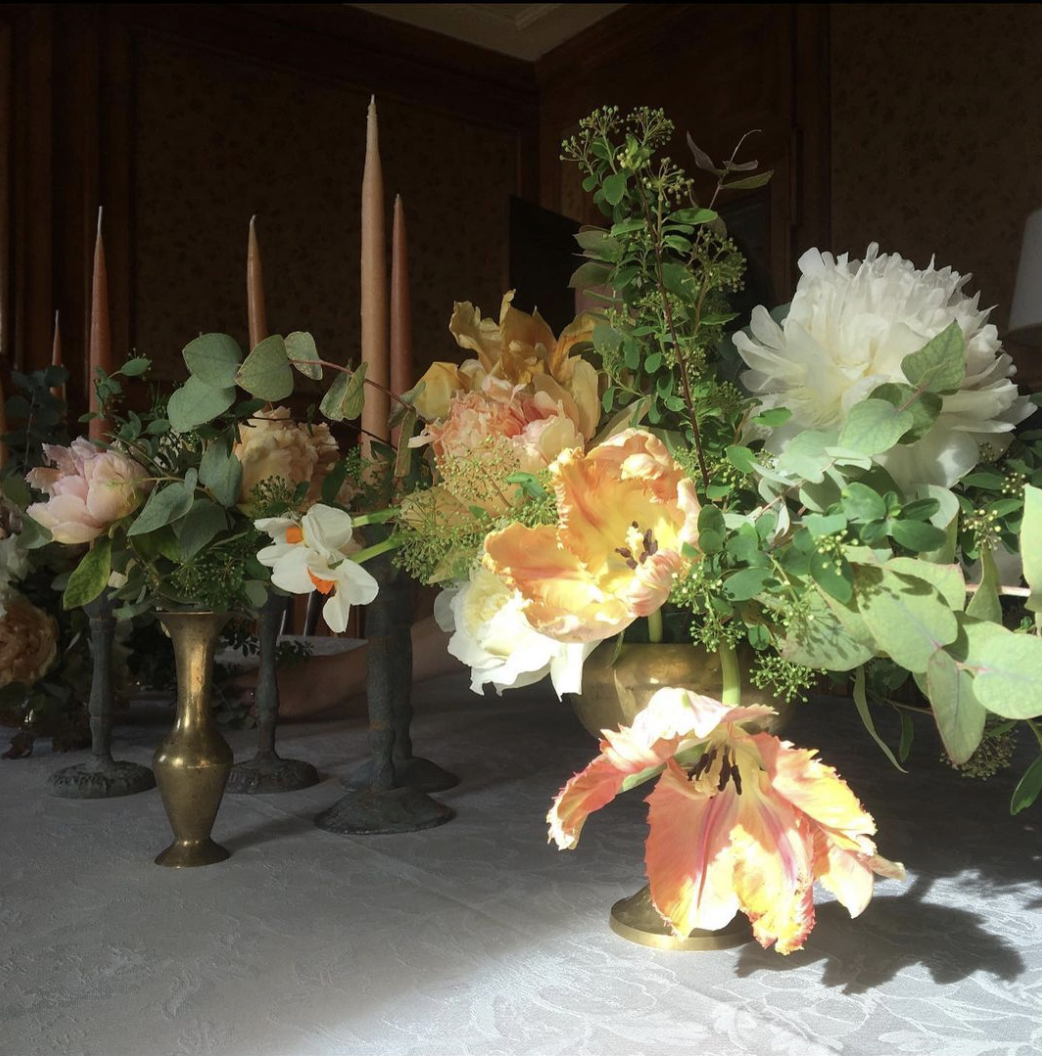 A floral arrangement with various flowers, including a large white flower, orange lilies, and pink peonies, displayed in brass vases on a table with tall pink candles in the background.