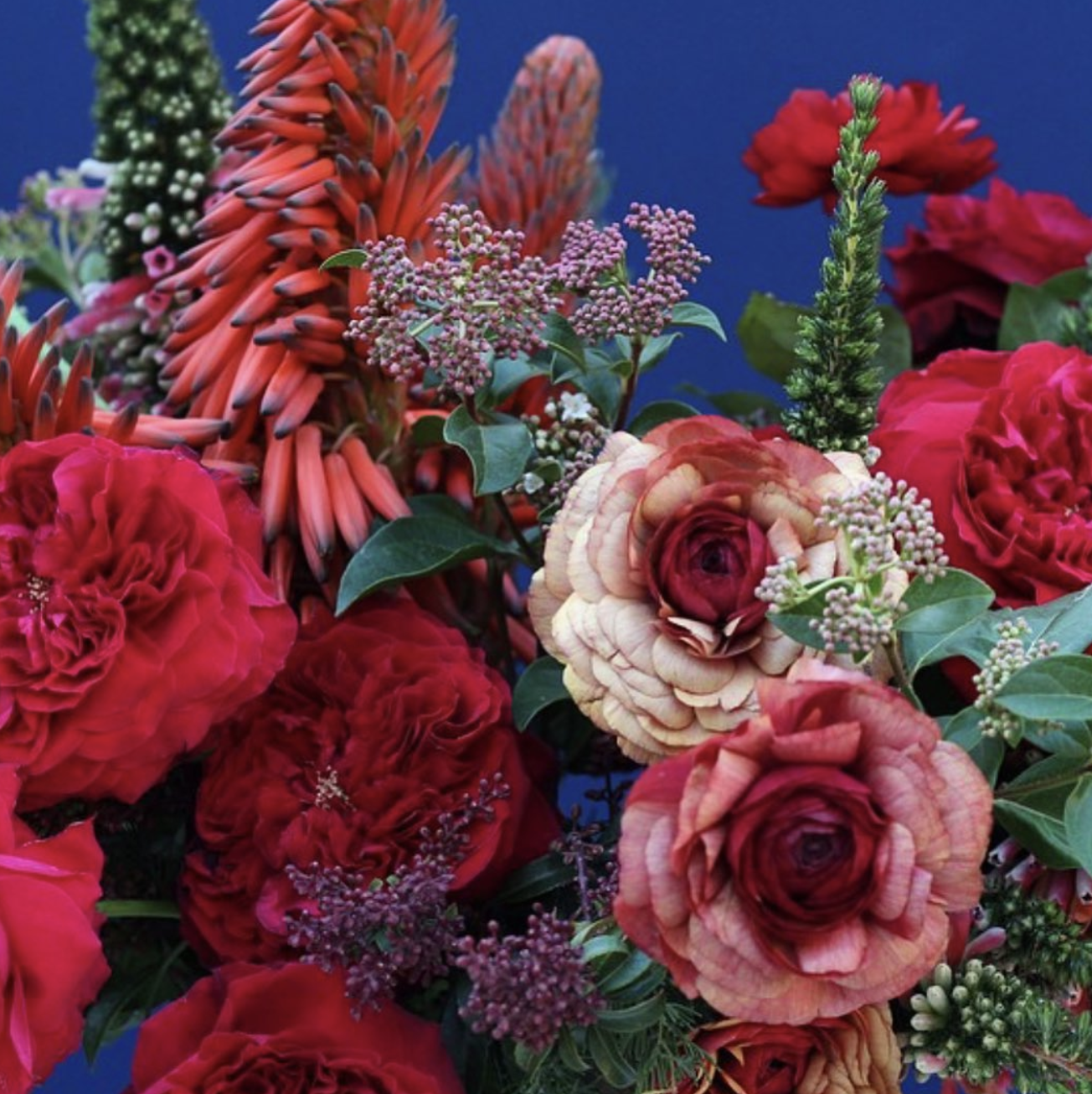 A vibrant bouquet of assorted red and pink flowers including roses, lilies, and other blossoms with green leaves against a dark background.