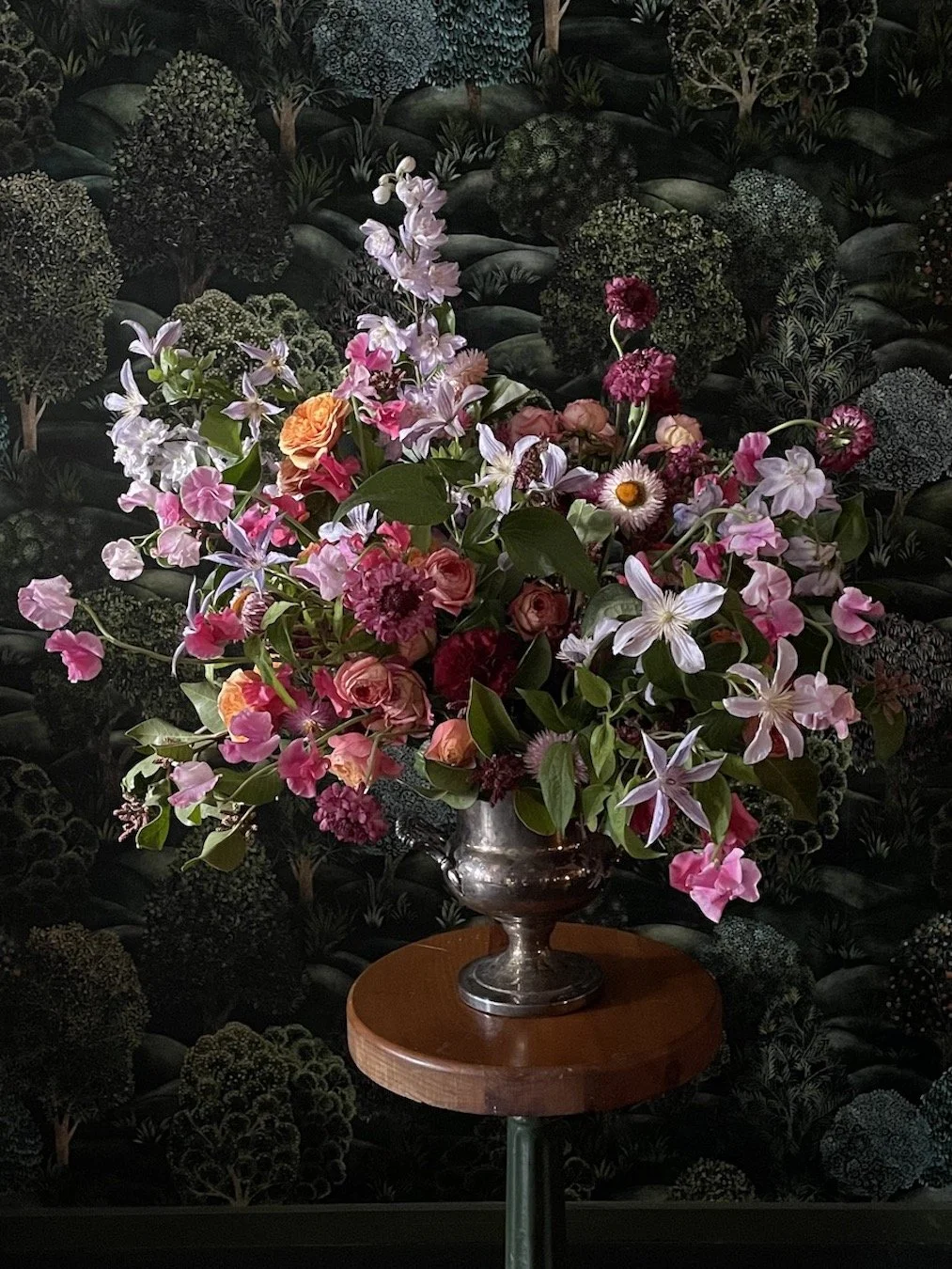 A floral arrangement in a silver vase displayed on a small round wooden table against a dark, patterned background.