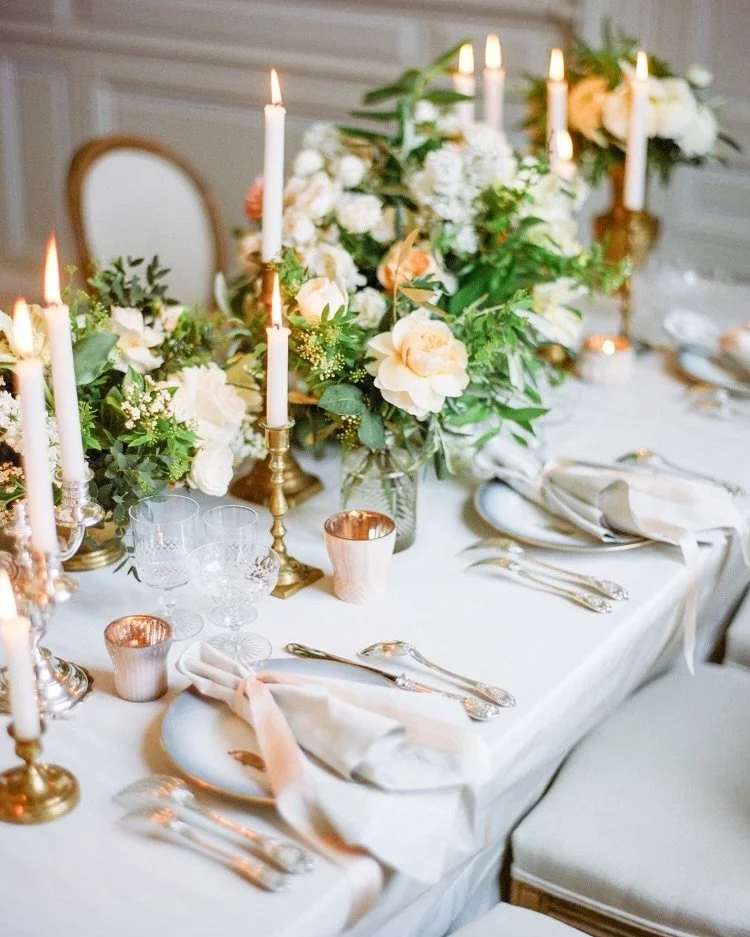 Elegant dining table decorated with white flowers, green foliage, gold candle holders, lit candles, and glassware.