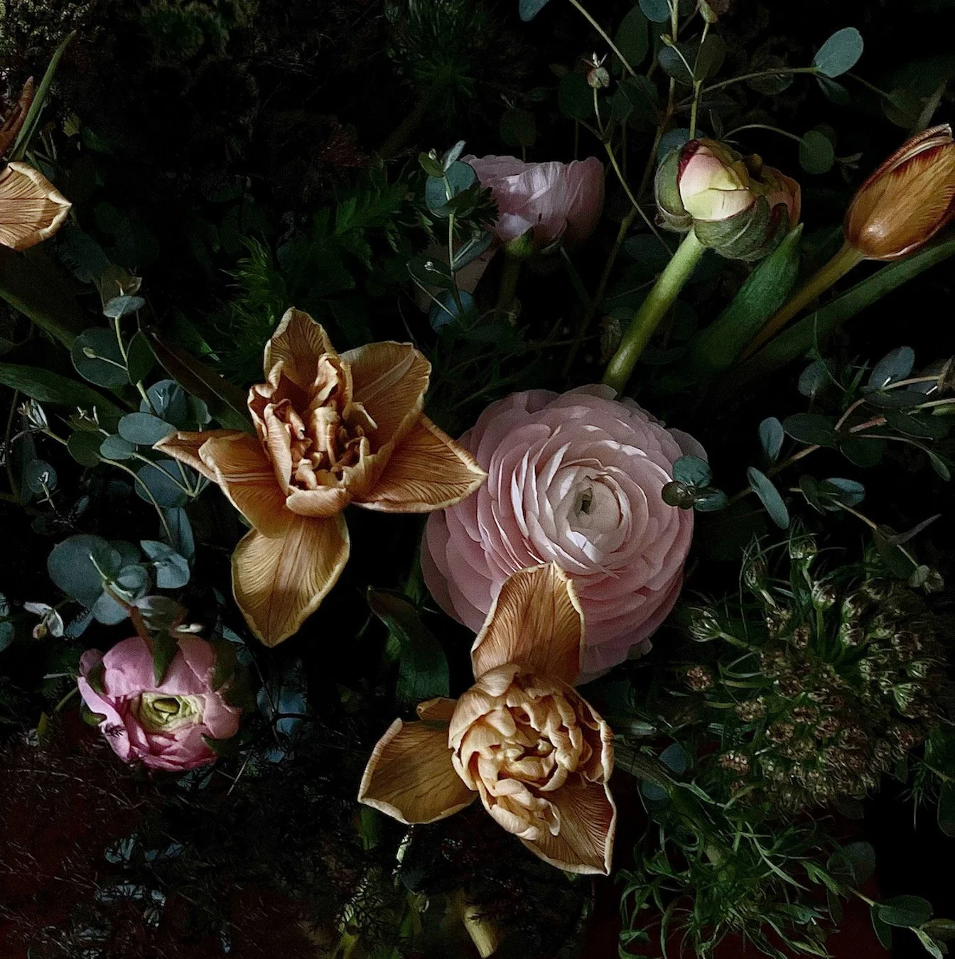 Close-up of various flowers, including pink roses and yellow tulips, surrounded by green leaves and foliage in a garden setting.