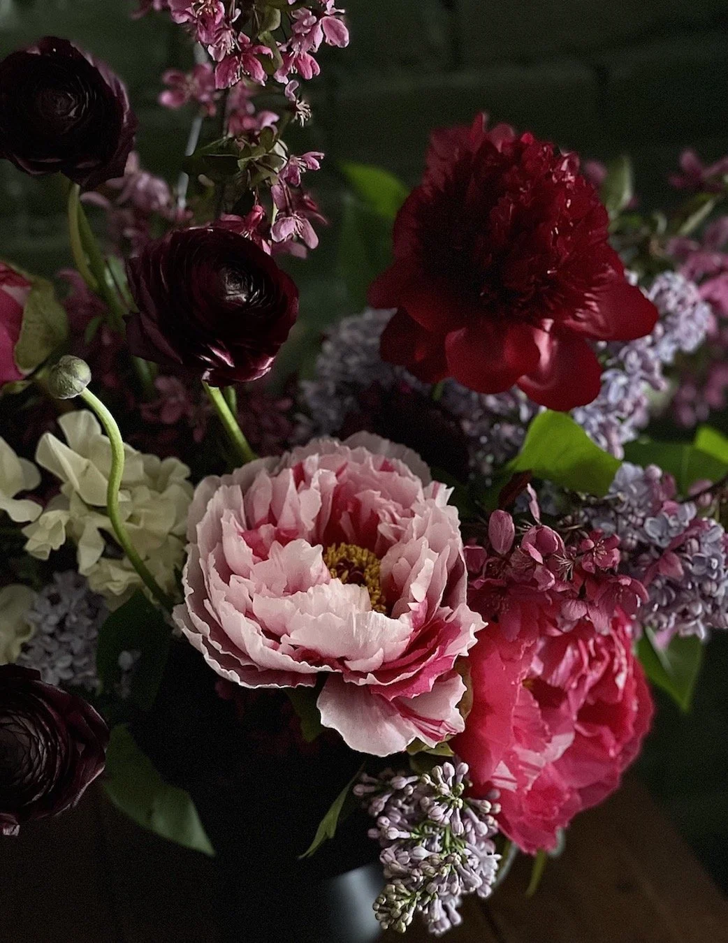 A bouquet of various pink, red, and dark purple flowers, including peonies, ranunculus, and lilacs, with green leaves in a dark vase.