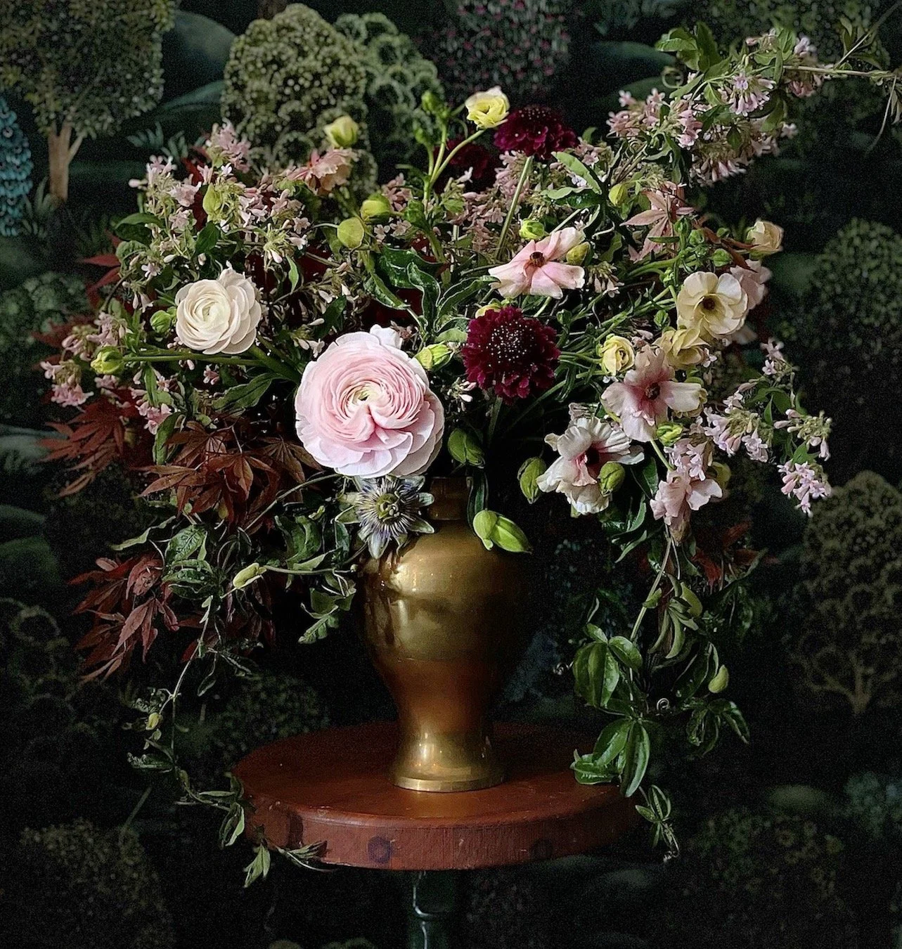 A bouquet of mixed flowers in a gold vase on a small round wooden table, with a dark, leafy background.