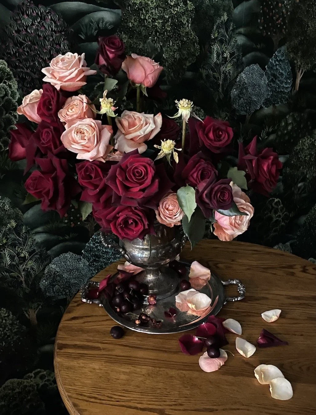 A bouquet of pink, red, and peach roses in a silver vase on a wooden table, with fallen rose petals and dark berries on a silver tray, set against a lush green foliage background.