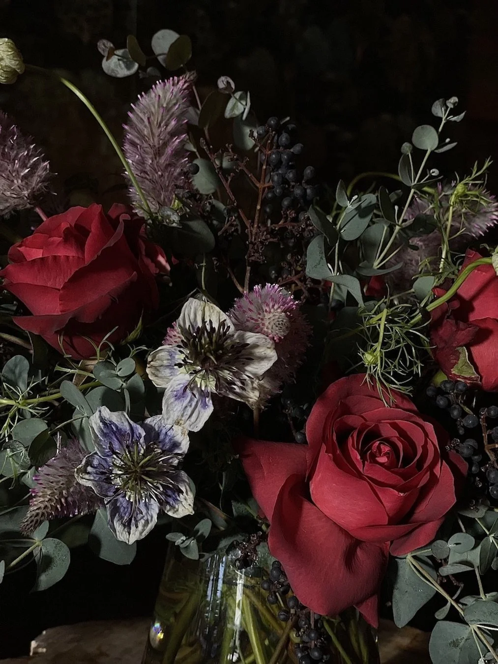 A bouquet of red roses, white and purple flowers, green leaves, and small dark berries in a clear glass vase.