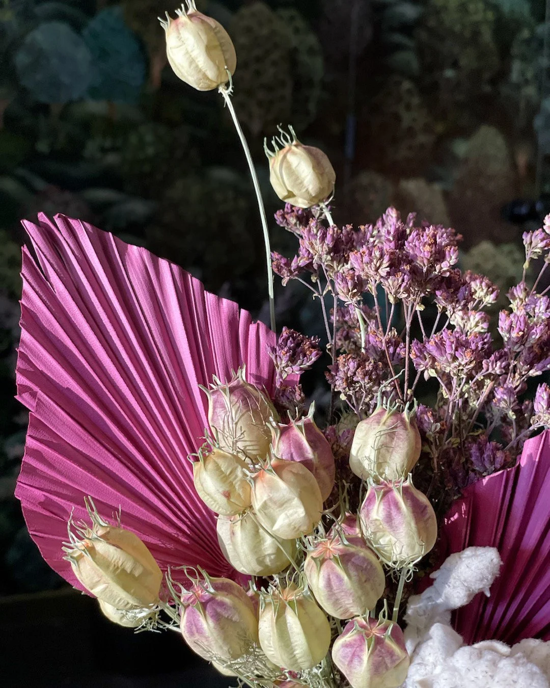 A floral arrangement with pink fan-shaped leaves, purple flowers, and dried seed pods, set against a dark background.