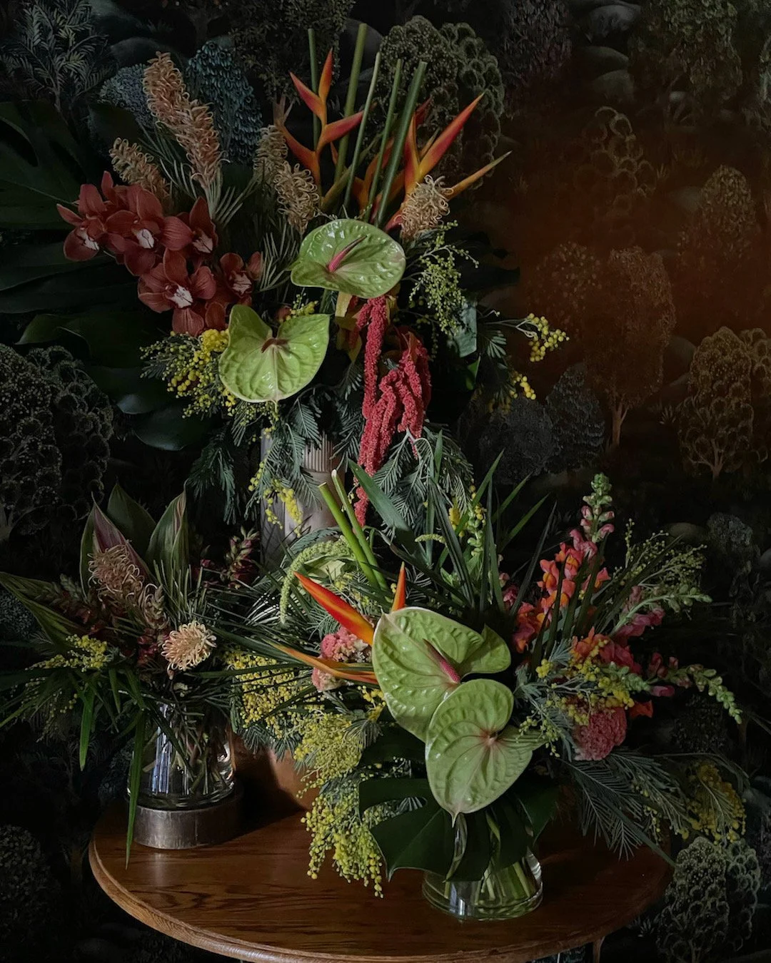 Two floral arrangements in vases with tropical flowers and green foliage, placed on a wooden table.