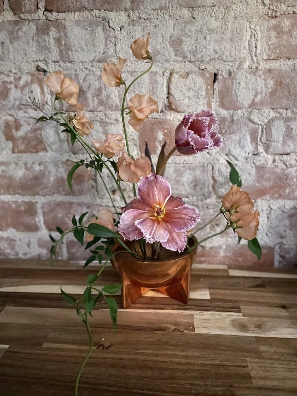 Pink and peach artificial flowers arranged in a small amber glass vase on a wooden surface against an exposed brick wall background.