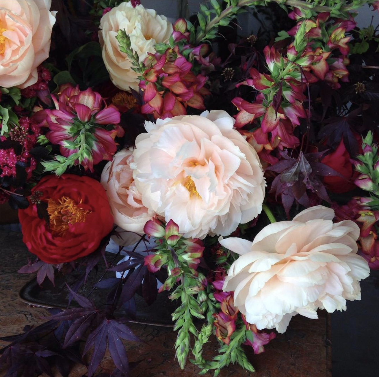 A colorful arrangement of peonies and other flowers with cream, pink, red, and purple blossoms and green and purple foliage.
