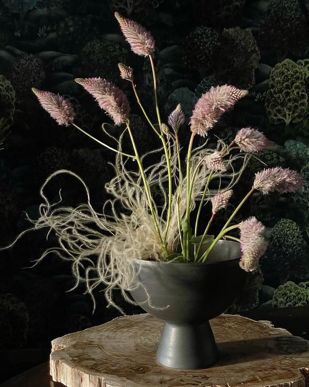 A black ceramic vase with pinkish plumes and curly, wispy white foliage inside, placed on a wooden slab against a dark background with green foliage patterns.
