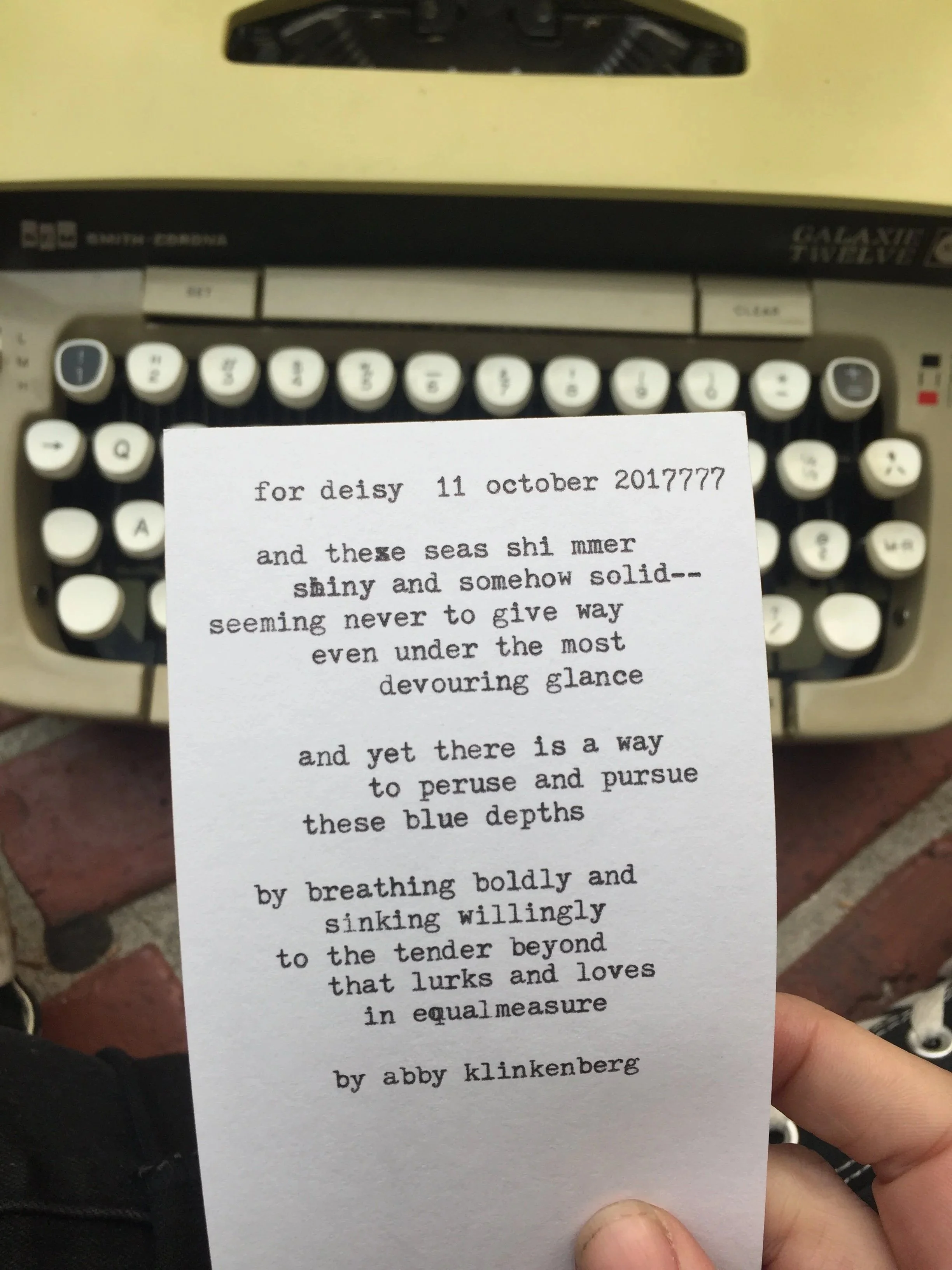 A close-up of a typed poem on a small piece of paper, held by a person's hand, with a vintage typewriter in the background.