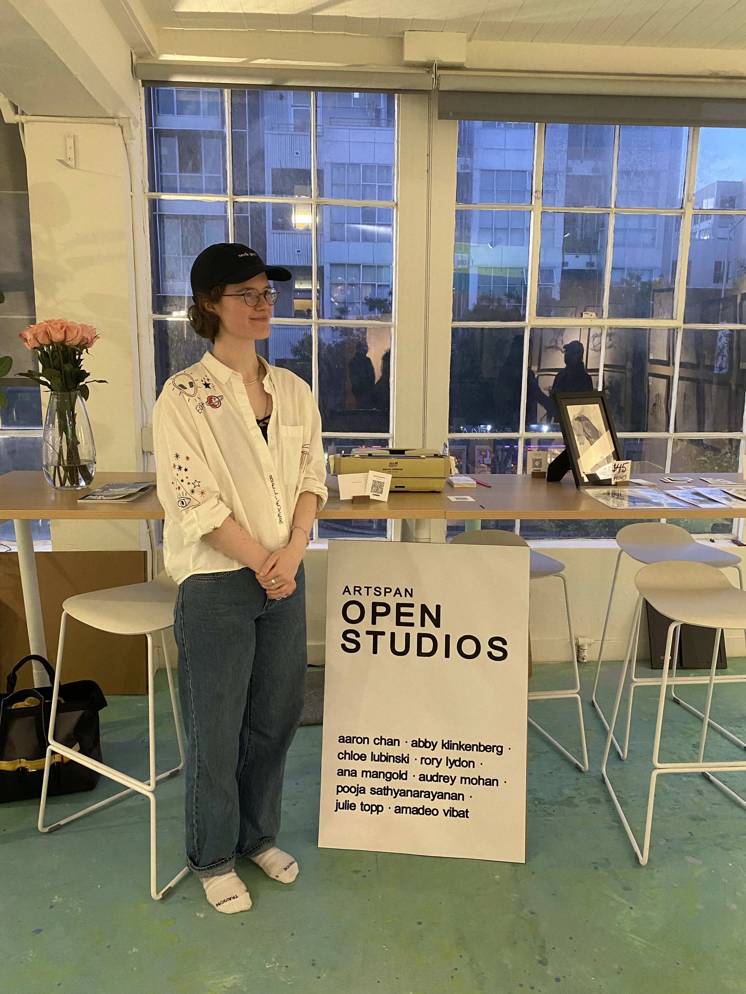 A woman with glasses and a black baseball cap standing next to a white sign that reads "ARTSPAN OPEN STUDIOS" with a list of artists' names. There is a table with items on it, a vase with pink roses, and a large window showing a cityscape outside.
