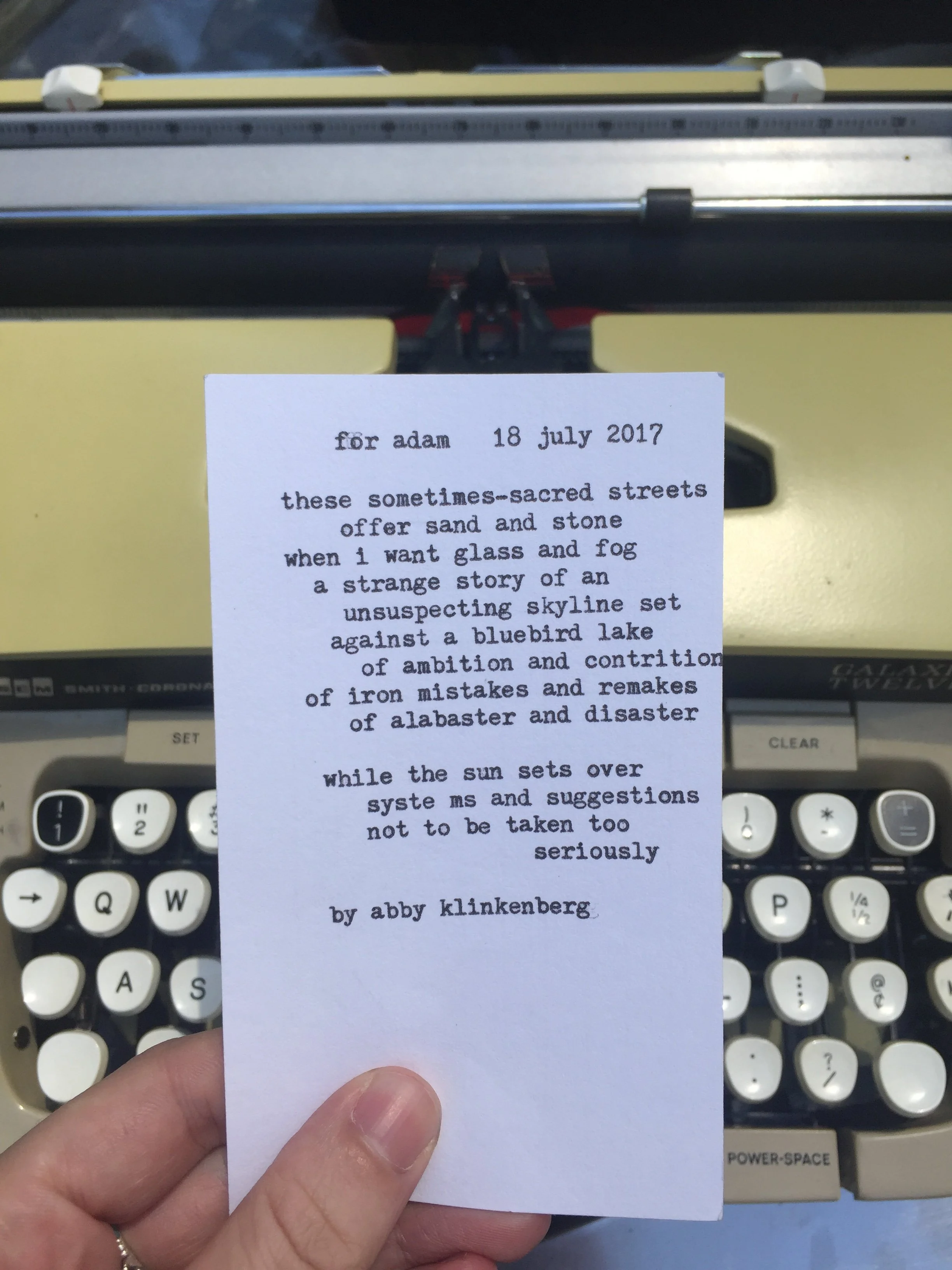 A person's hand holding a handwritten note in front of a vintage typewriter. The note contains a poetic message.