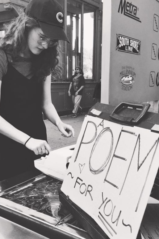 A young woman with glasses and curly hair, wearing a baseball cap and a black top, stands at a table with a sign that says "POEM FOR YOU" and a small heart drawing. She appears to be preparing or selling poetry, with a background featuring a Metra sign and South Shore Line signage at a train station.