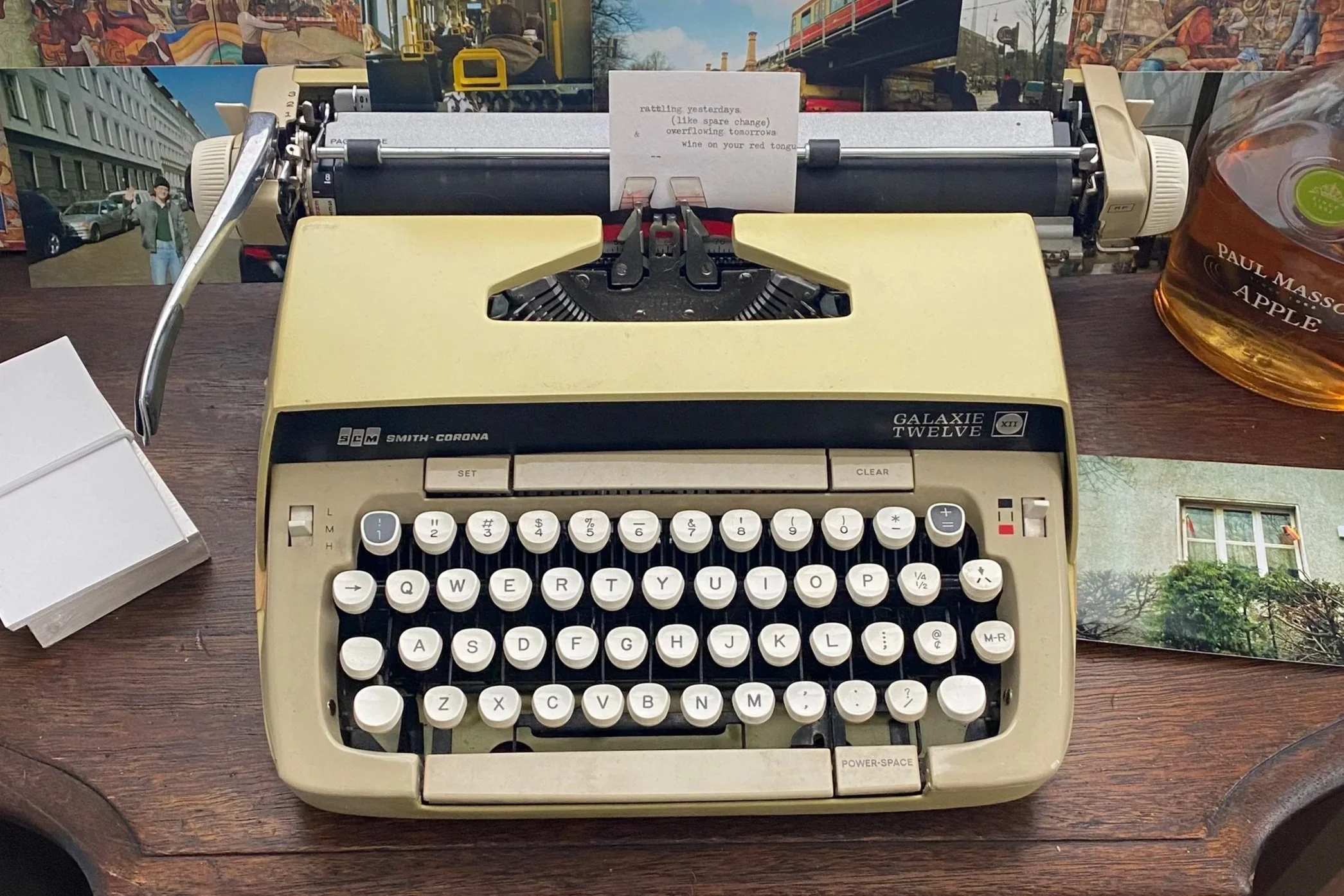 A vintage beige typewriter on a wooden table with a white sheet of paper inserted, a jar of maple apple syrup on the right, and a colorful outdoor city street visible through the window in the background.