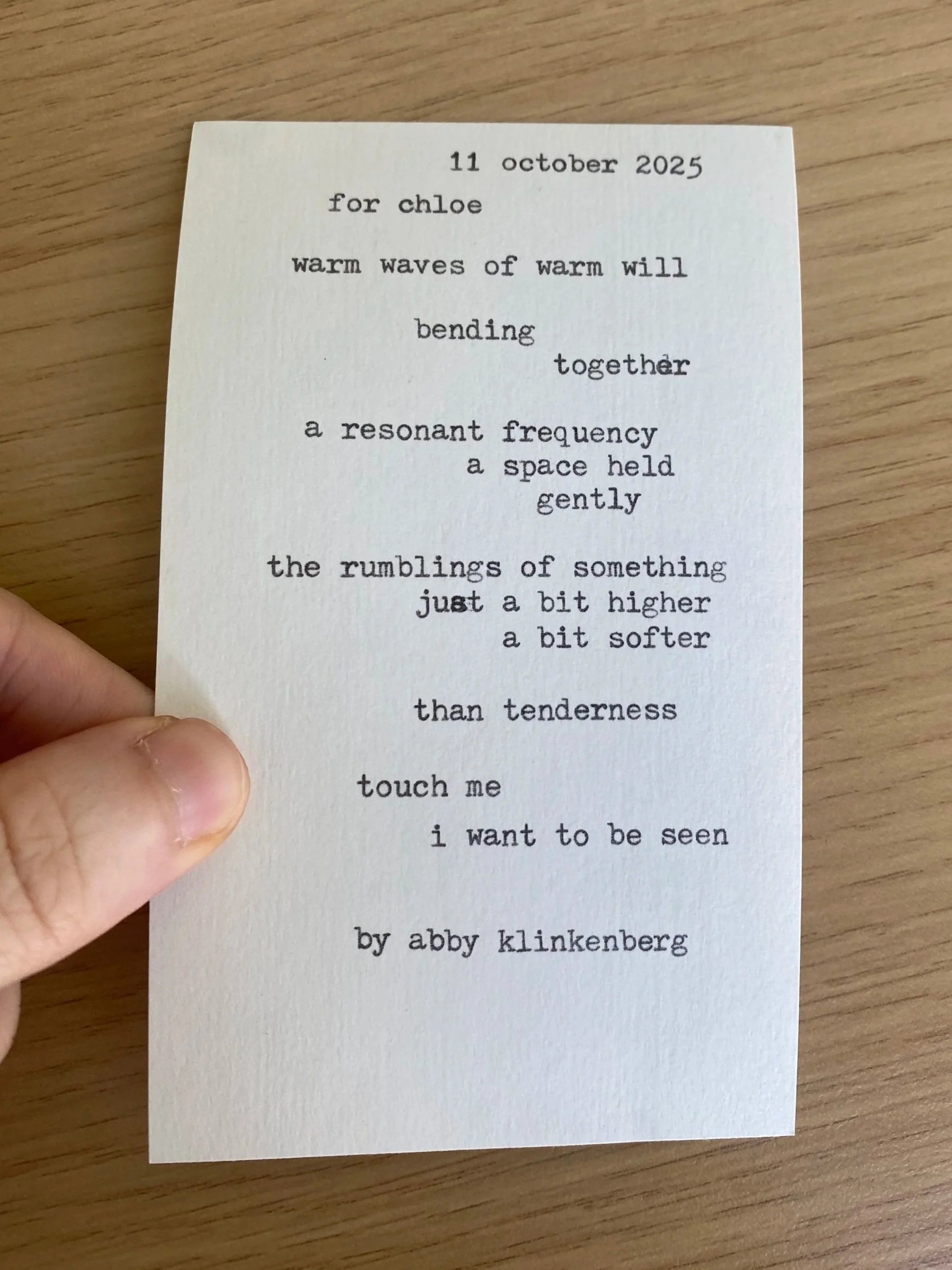 A poem written on a white piece of paper with typewriter-style font, held by a person's hand, placed on a wooden surface.