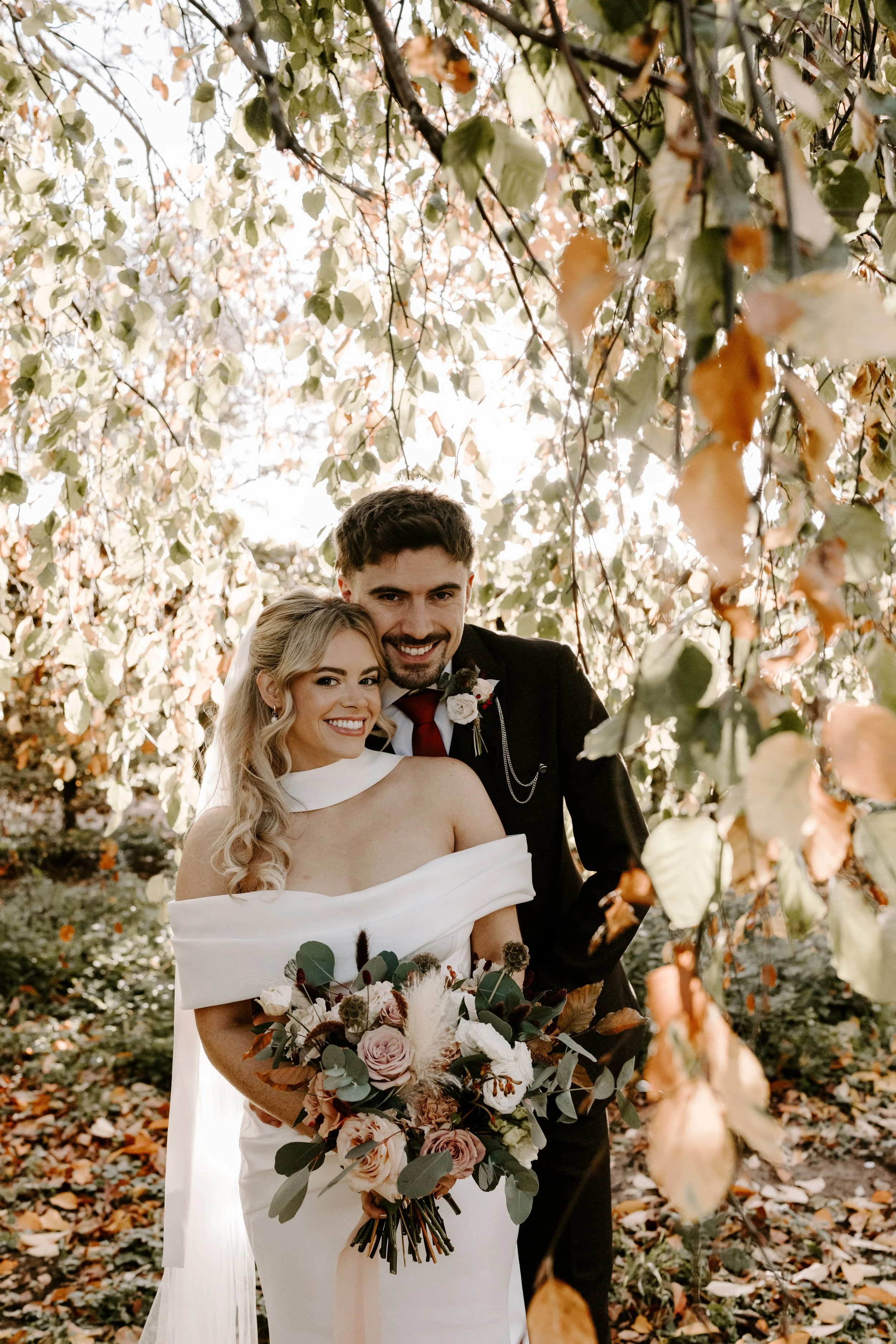 A smiling bride and groom pose outdoors among autumn leaves, with the bride holding a bouquet of roses, greenery, and dried flowers.