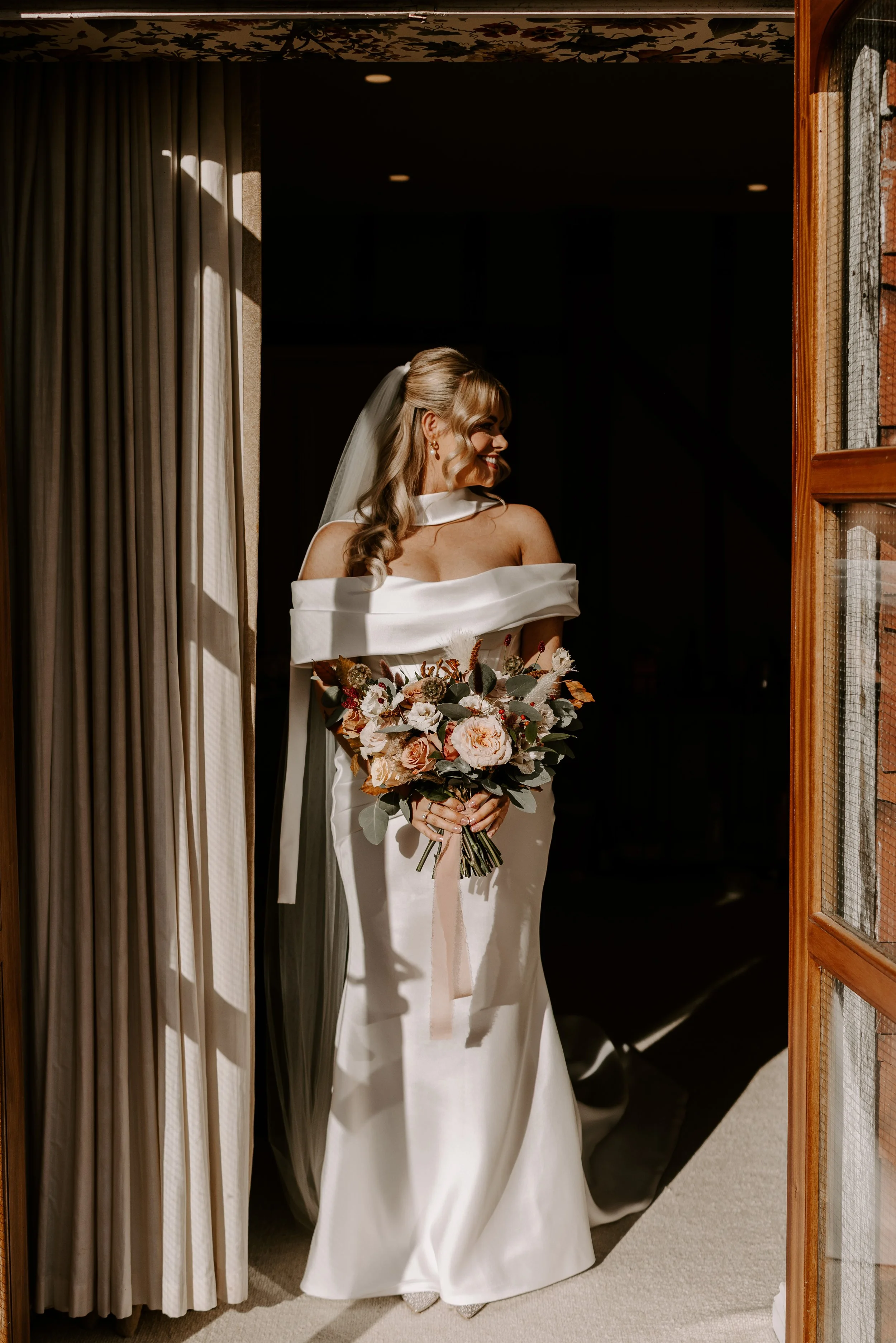 A bride in a white off-shoulder wedding dress holding a bouquet of flowers, standing in a doorway with sunlight casting shadows on her face and dress.