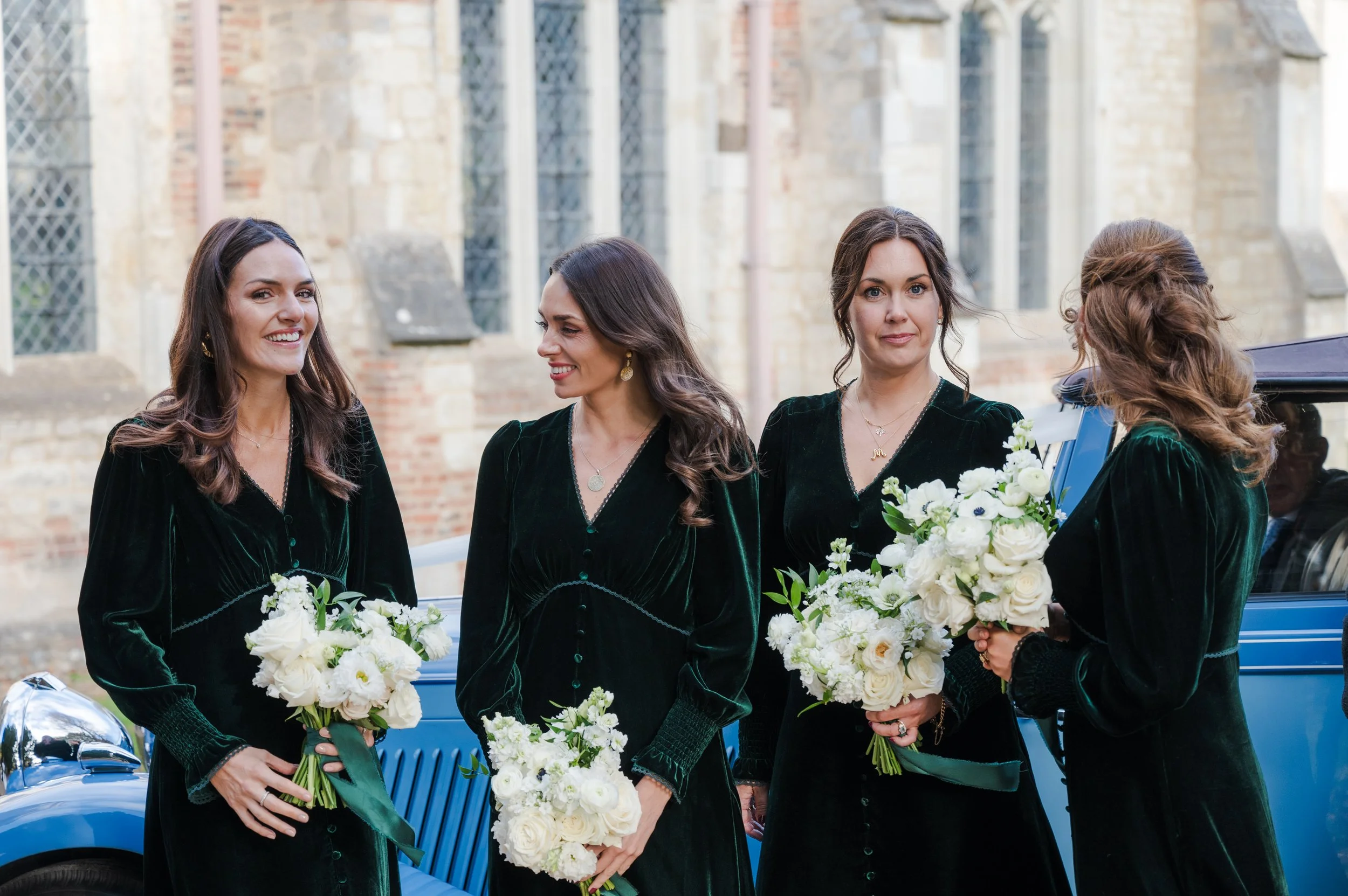 Four women dressed in black velvet dresses holding white floral bouquets, standing outdoors near a vintage blue car with a brick building in the background.