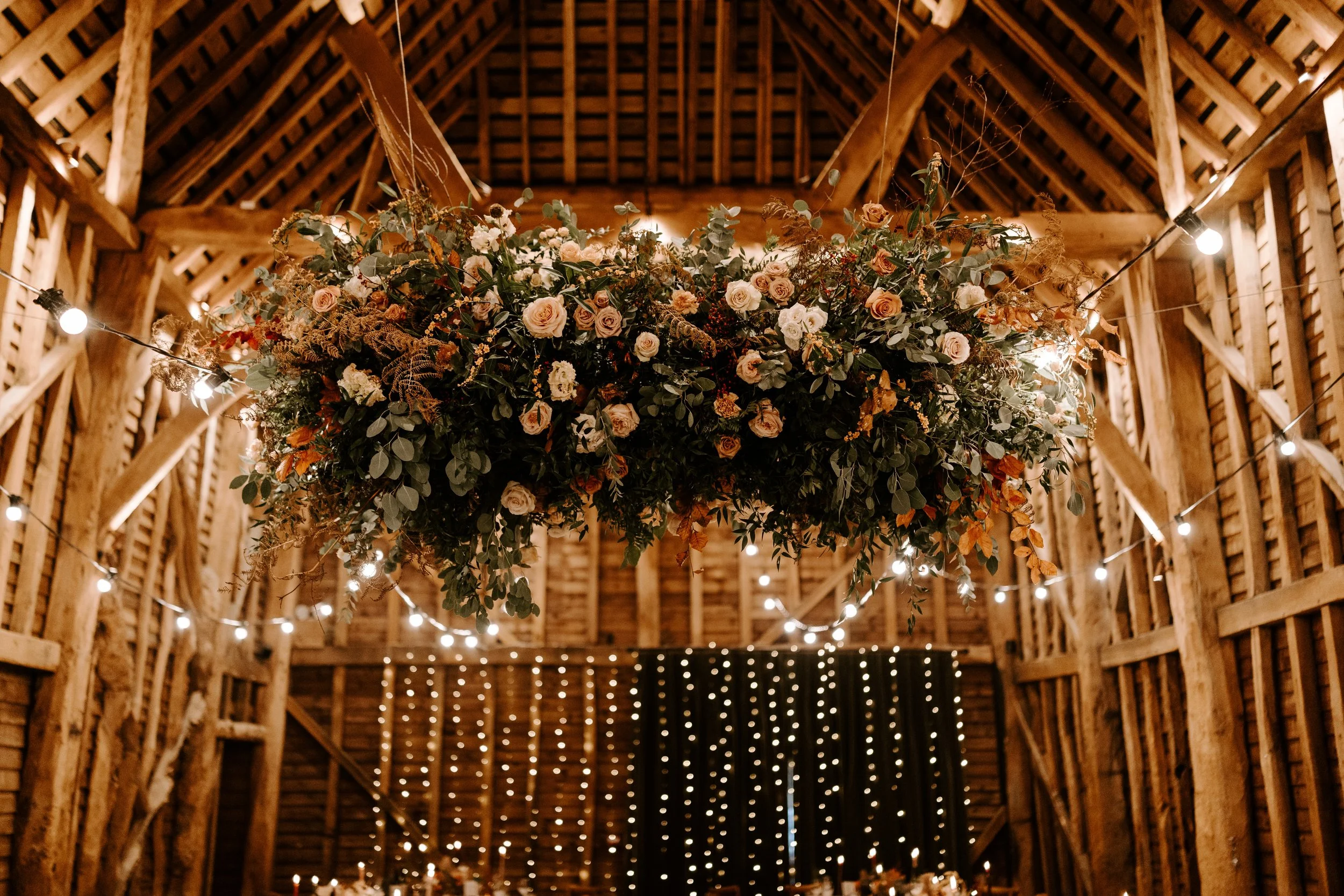 A large floral arrangement with pink and white roses, greenery, and dried foliage, suspended from the ceiling of a rustic wooden barn decorated with string lights.