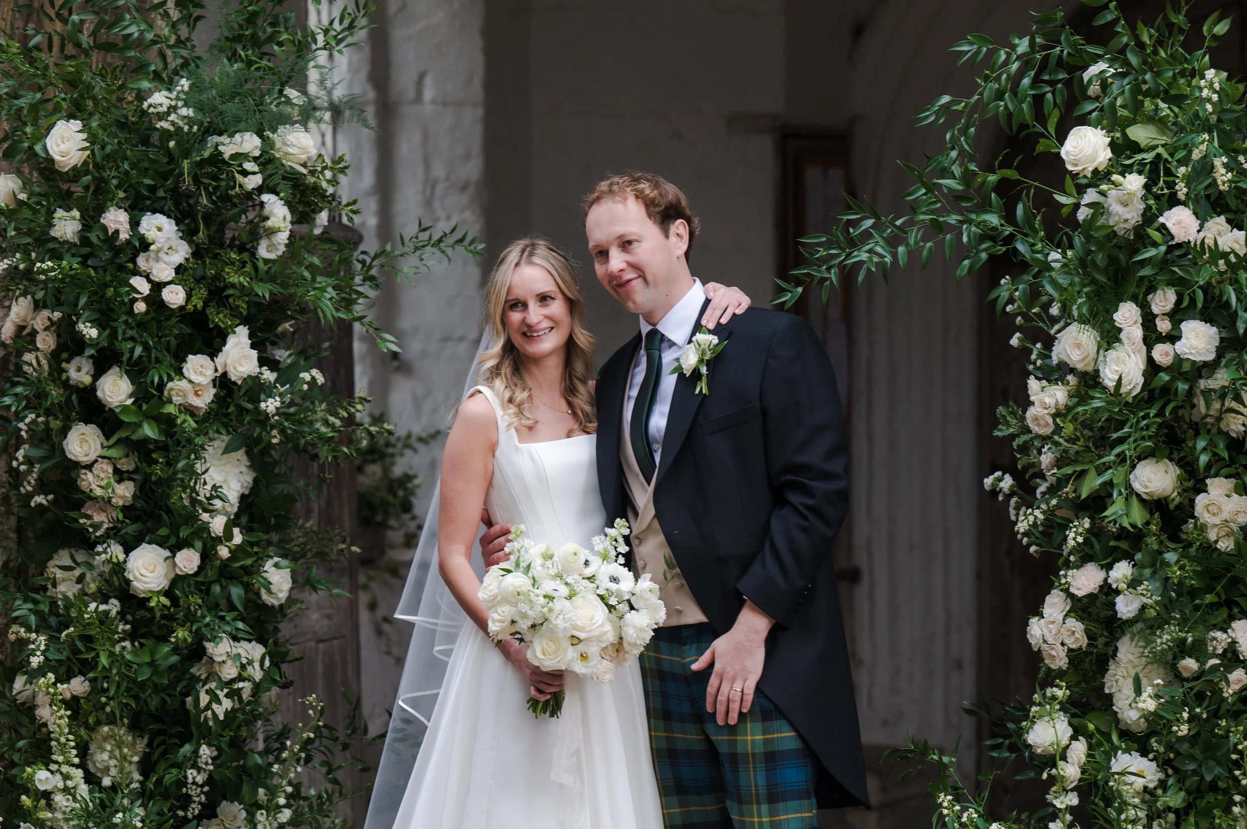 A bride and groom posing together on their wedding day, surrounded by white and green floral decorations.