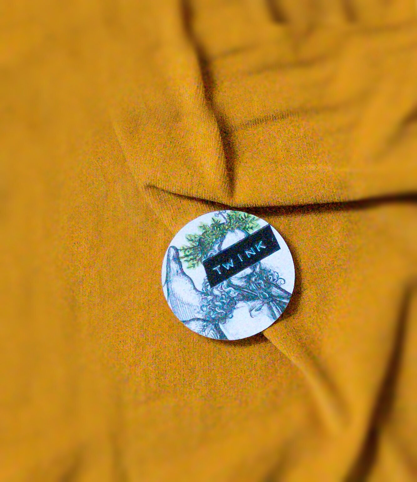 Circular sticker with glitchy black and white engraving of an angel, his halo highlighted in yellow. The angel's face is obscured by a label reading "TWINK" in white embossing on black tape.