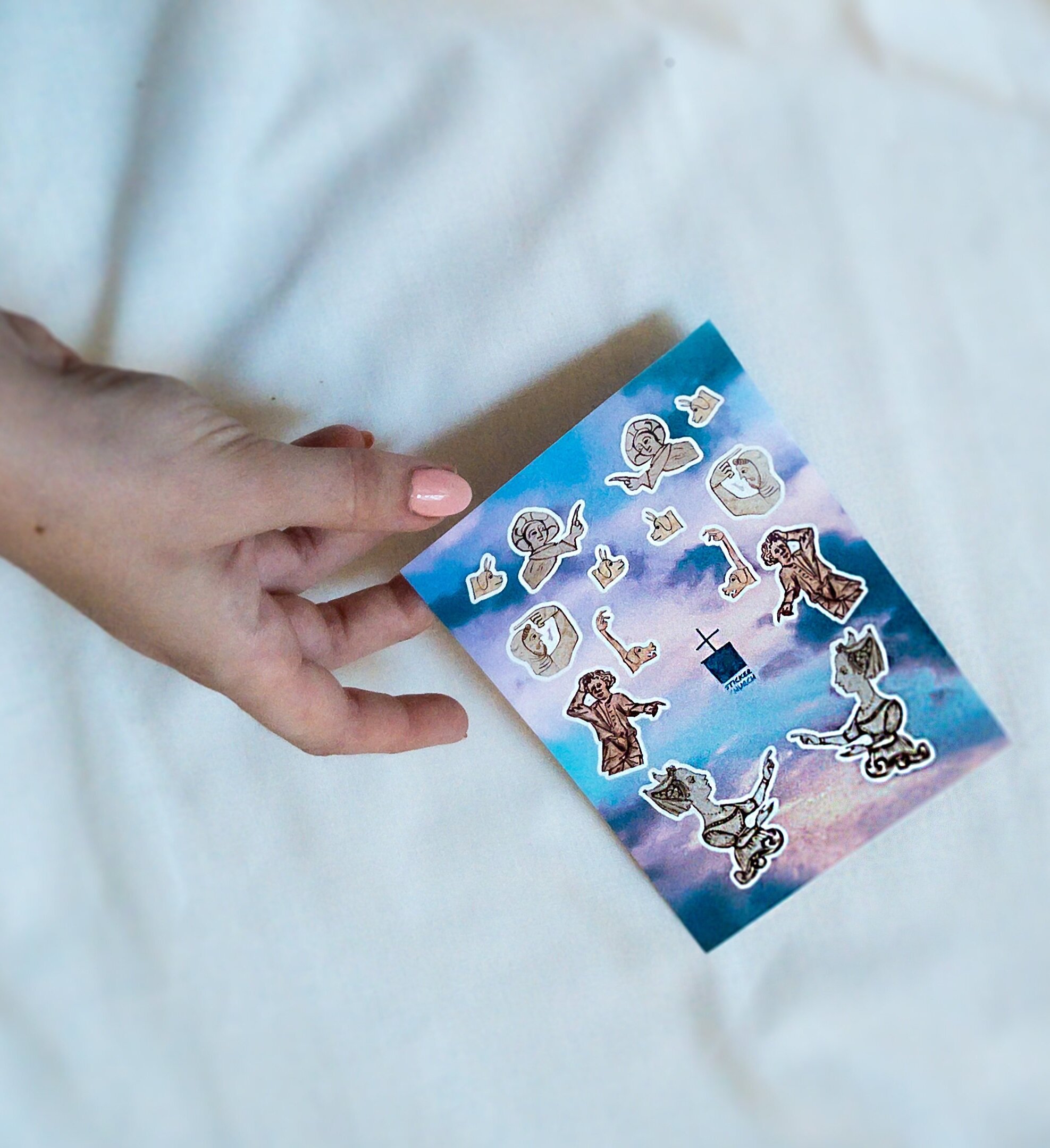 Hand with pale coral manicure holds a sticker sheet featuring designs of medieval manicules in the form of people and dogs, in front of a neutral background.